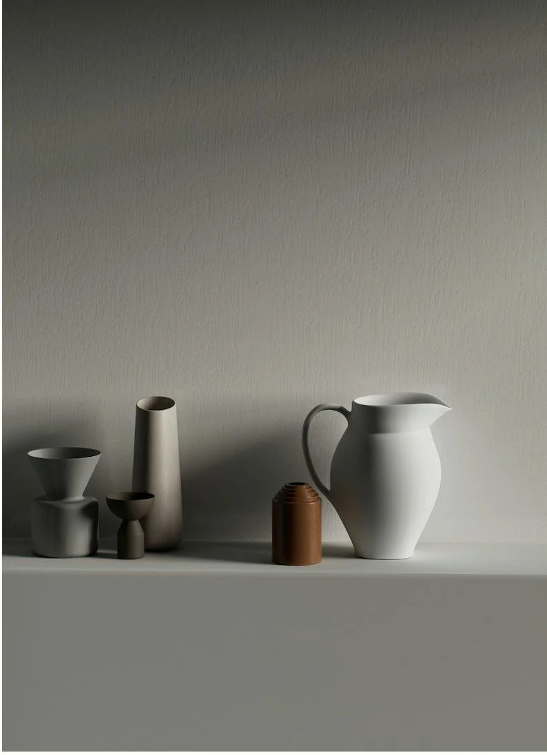 A minimalist arrangement of six ceramic vessels of varying shapes and sizes on a shelf against a neutral wall, including a large white pitcher, a small brown bottle, and several smaller vases.