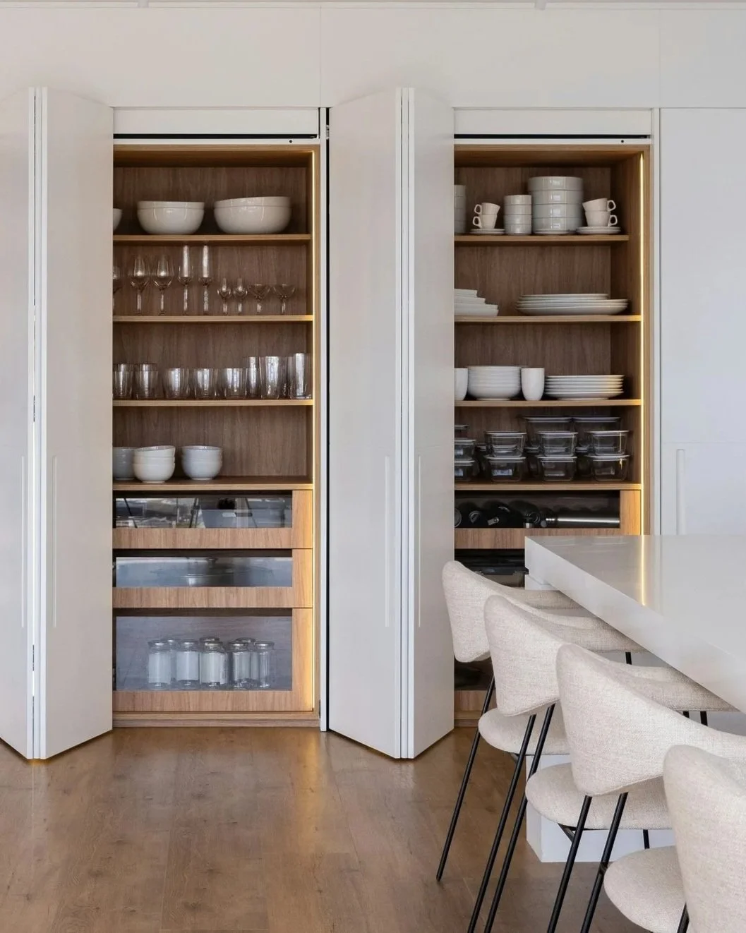 Open kitchen cabinets filled with white dishes, glassware, and stacking bowls, next to a white countertop with beige chairs.