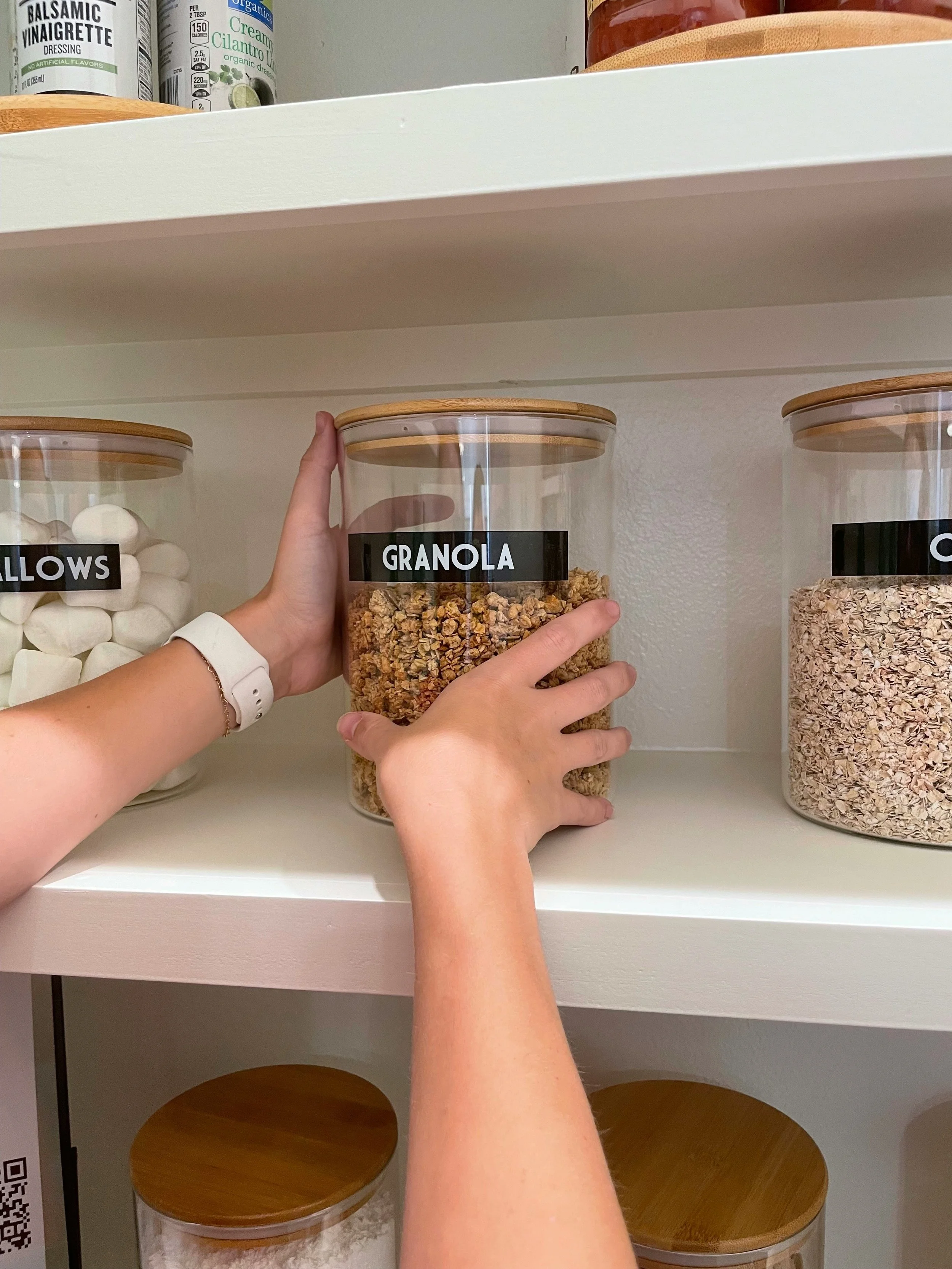 Person placing a jar labeled 'Granola' on a shelf in a pantry, with other jars labeled 'Marshmallows' and 'Oats' visible nearby.