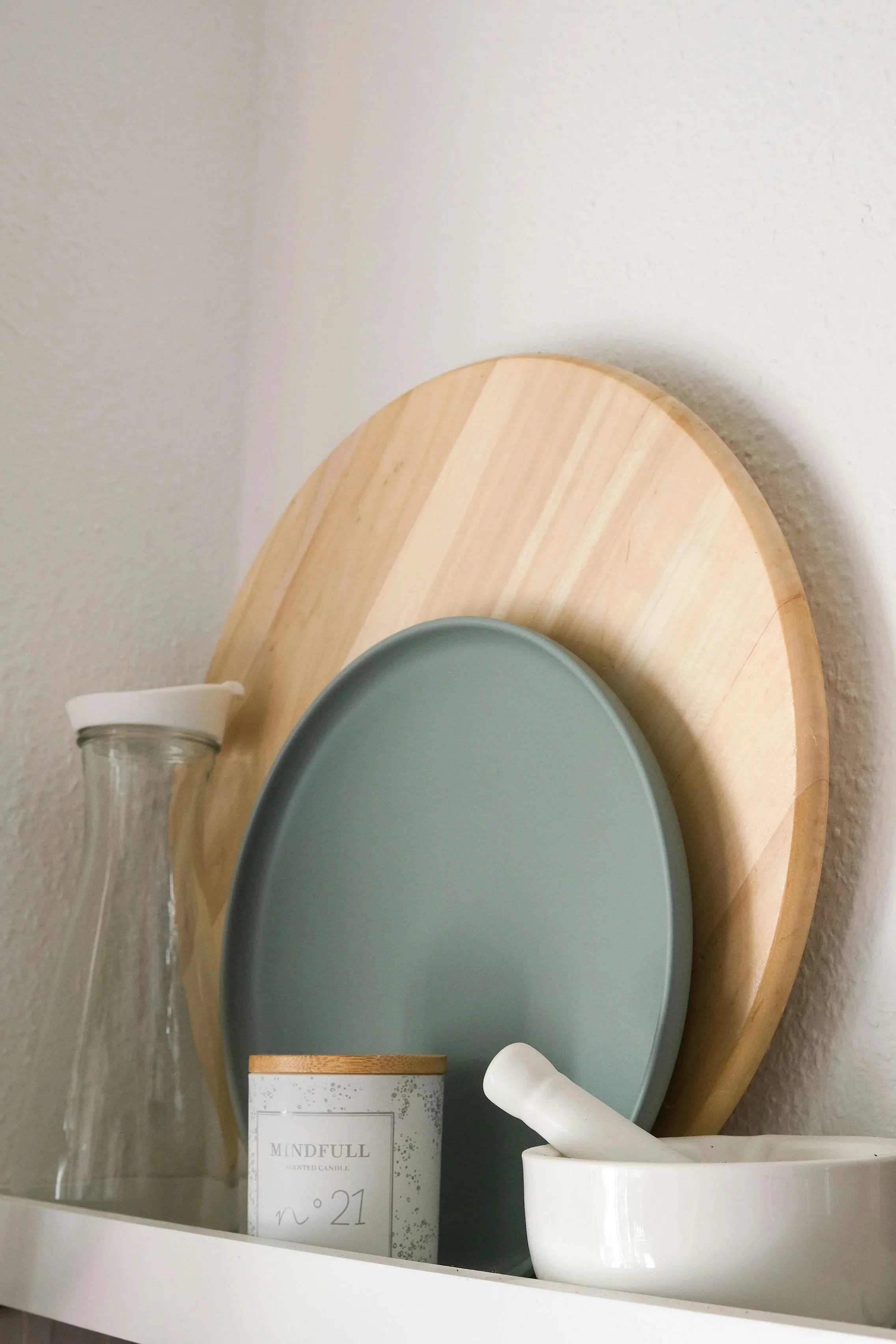 A small glass jar with a white lid, a box labeled 'Mindfull scented candle no. 21', a gray oval tray, a white bowl with a white mortar and pestle inside, and a white cup, all arranged on a round wooden tray against a white wall.