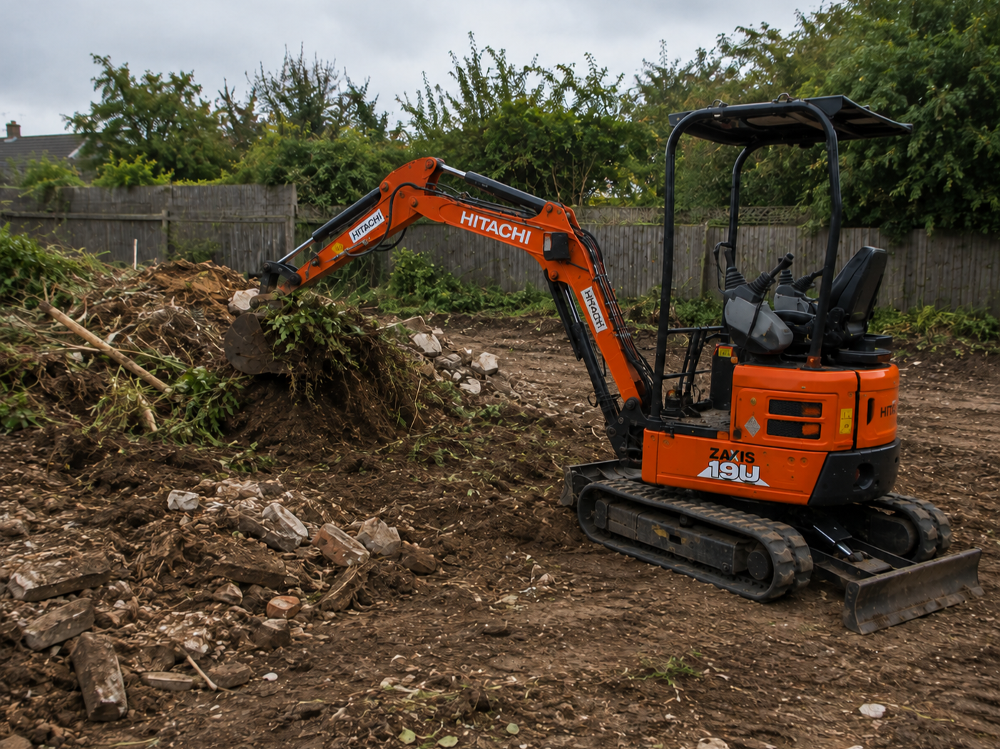 Small orange excavator working on construction or landscaping site, clearing dirt and debris, with a wooden fence and trees in the background.