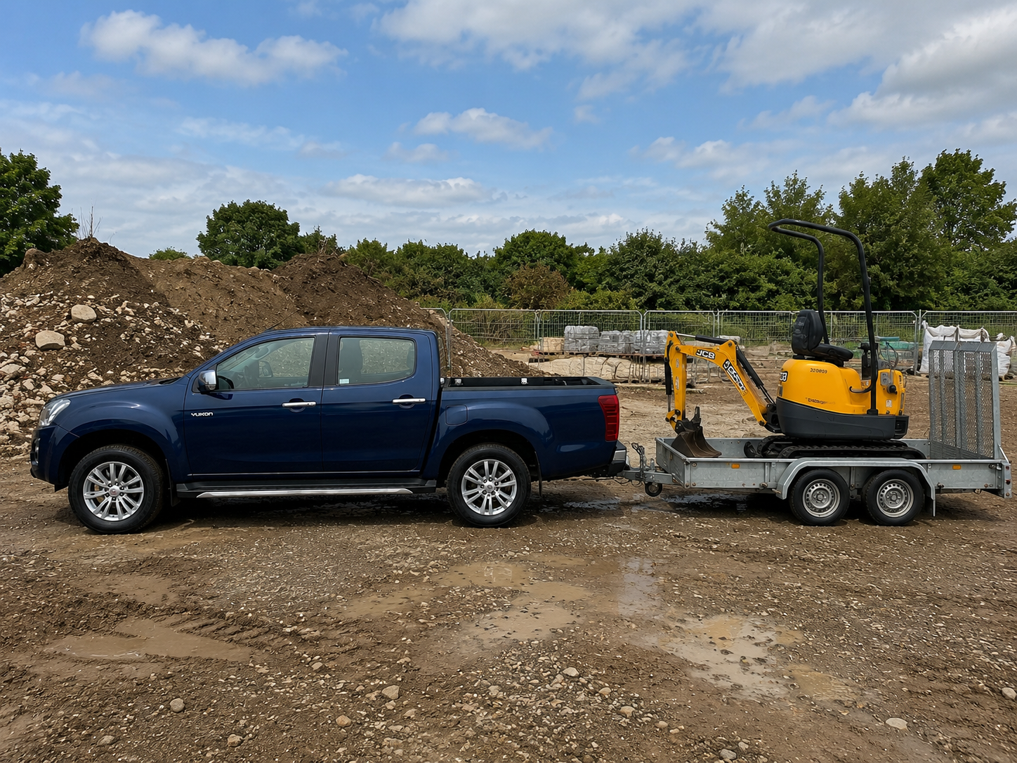 Blue pickup truck towing a small yellow JCB excavator on a construction site with dirt, rocks, and trees in the background.