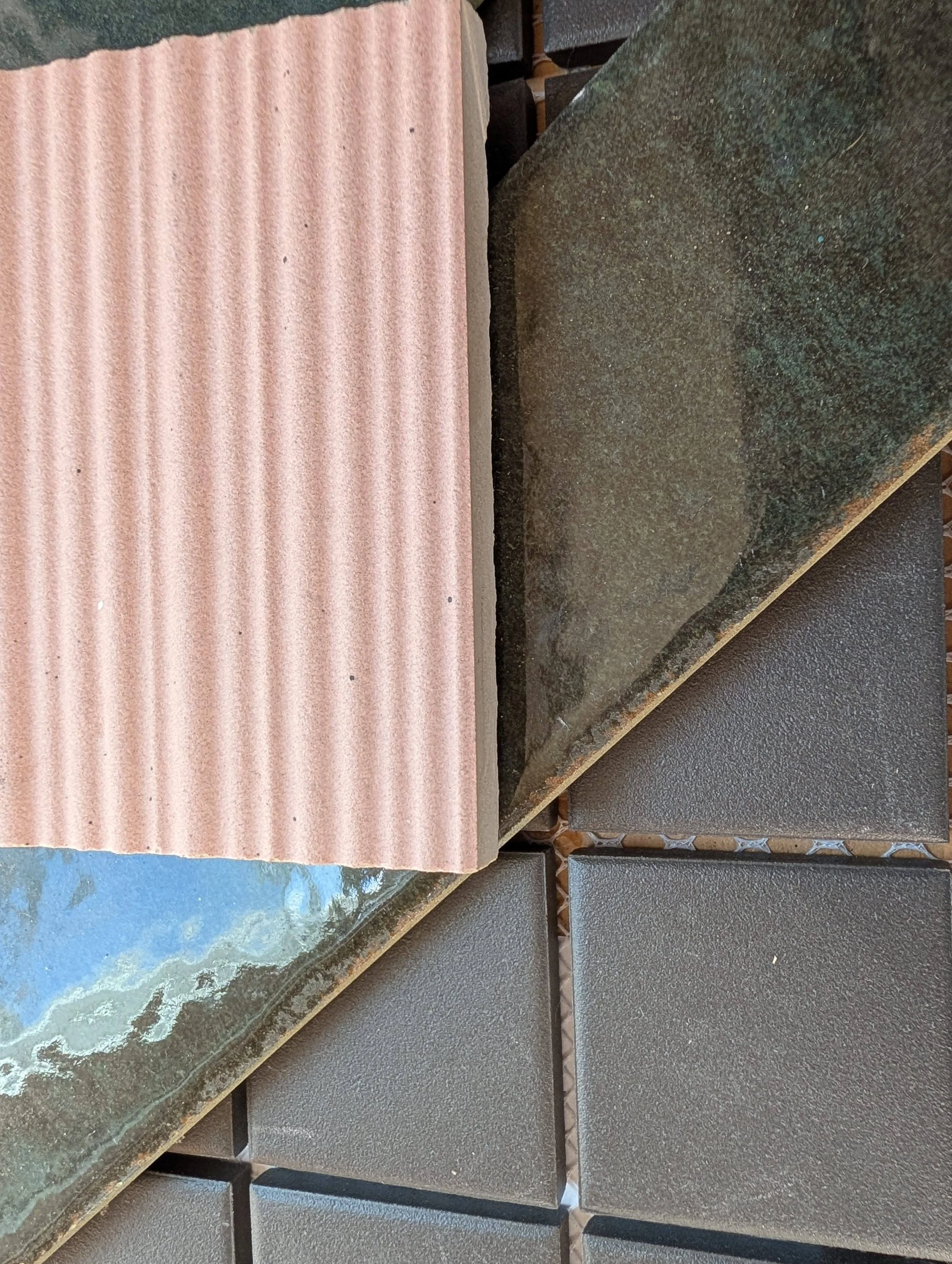 Close-up of variously colored and textured tiles, including pink corrugated tile, dark green rough tiles, and smooth black tiles, arranged in a pattern.