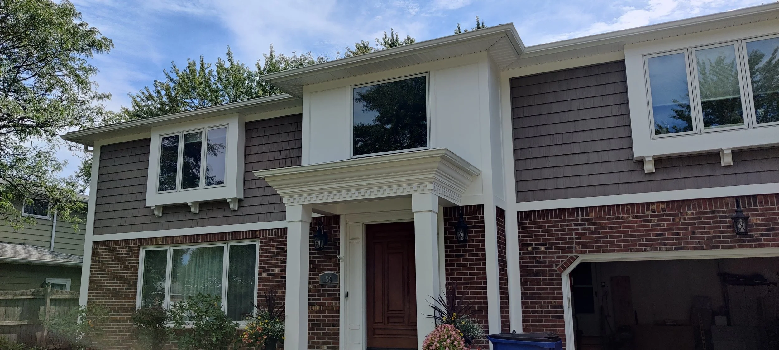 A two-story house with a brick lower level and dark gray shingle siding on the upper level. It has large white-framed windows, a covered front porch with decorative trim, and two wall-mounted lantern lights. There are trees in the background and a bl