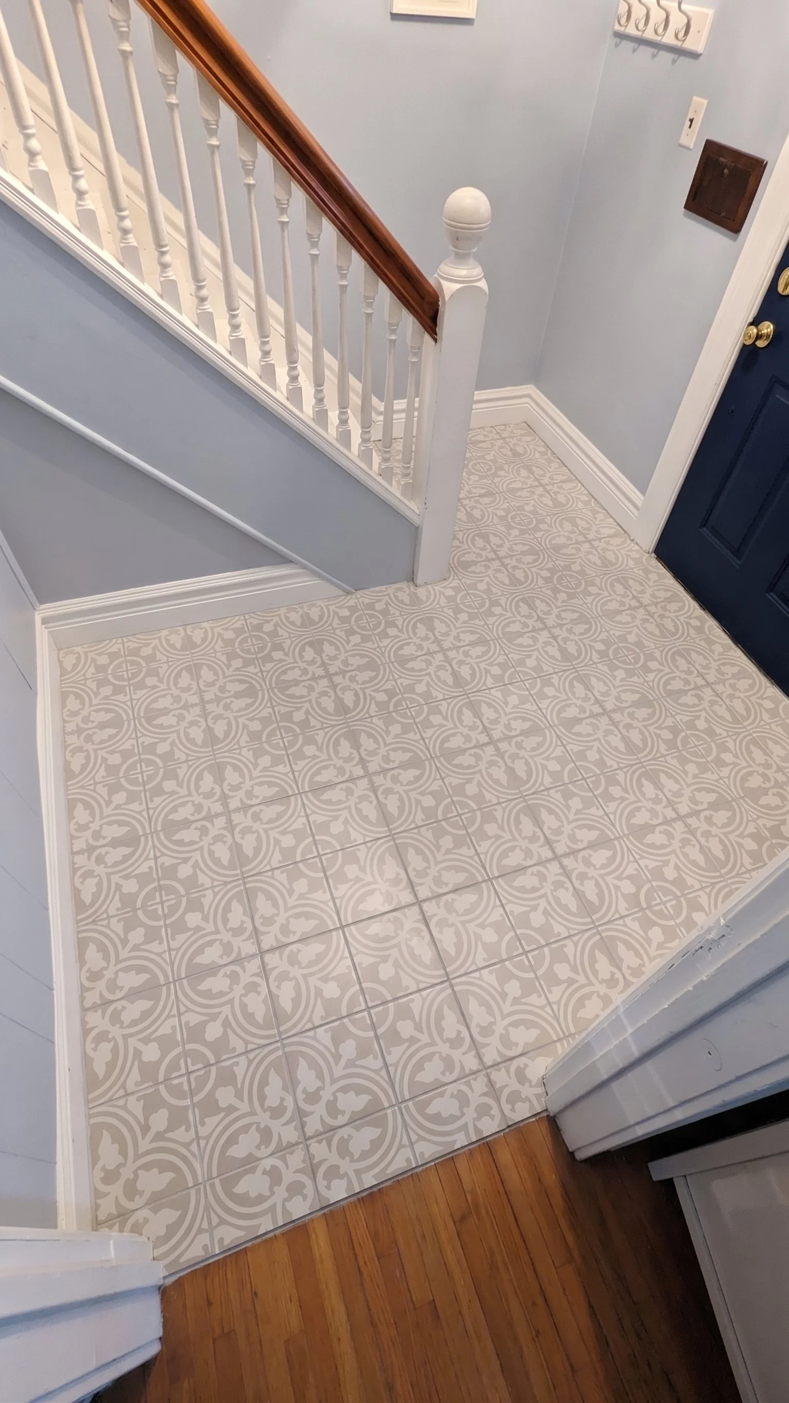 View of a small entryway with patterned ceramic tile flooring and hardwood flooring, white baseboards, staircase with white balusters and dark wood handrail, and a navy blue door.