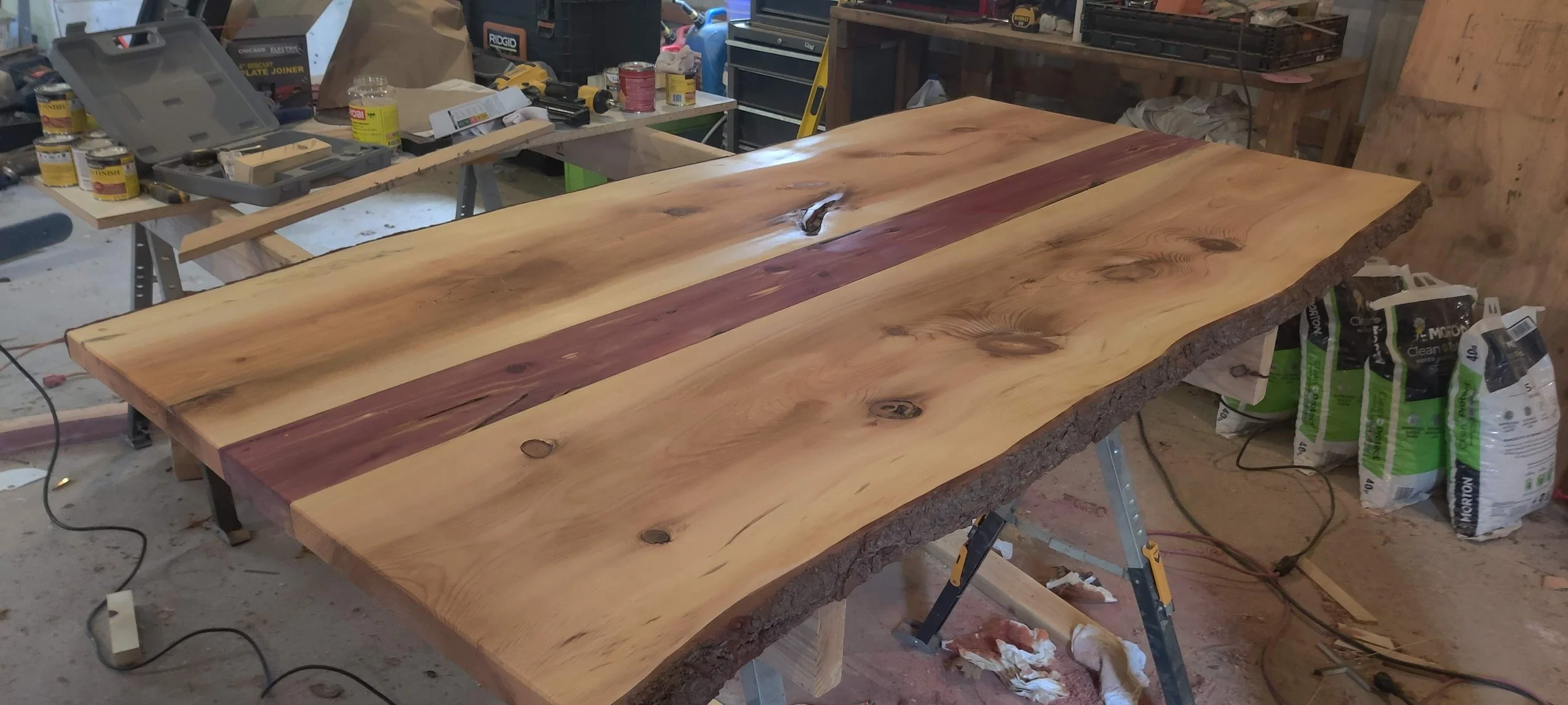 A large wooden table made from natural wood with live edges and a contrasting stripe of darker wood in the middle, in a woodworking workshop with tools, paint cans, and supplies around it.