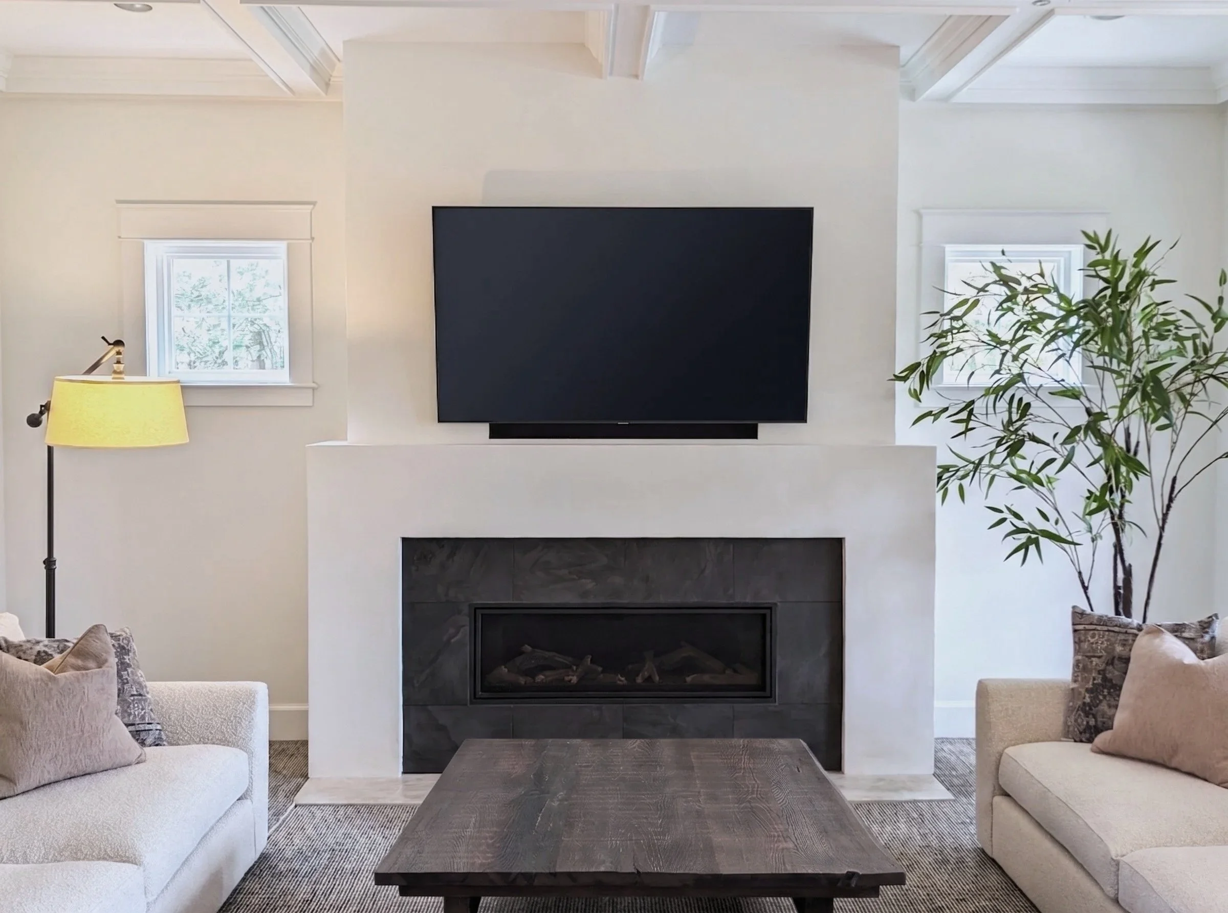 Living room with a black fireplace, white walls, two small windows, a tall yellow floor lamp, a large wooden coffee table, beige sofas, patterned pillows, and a green plant near the right window.