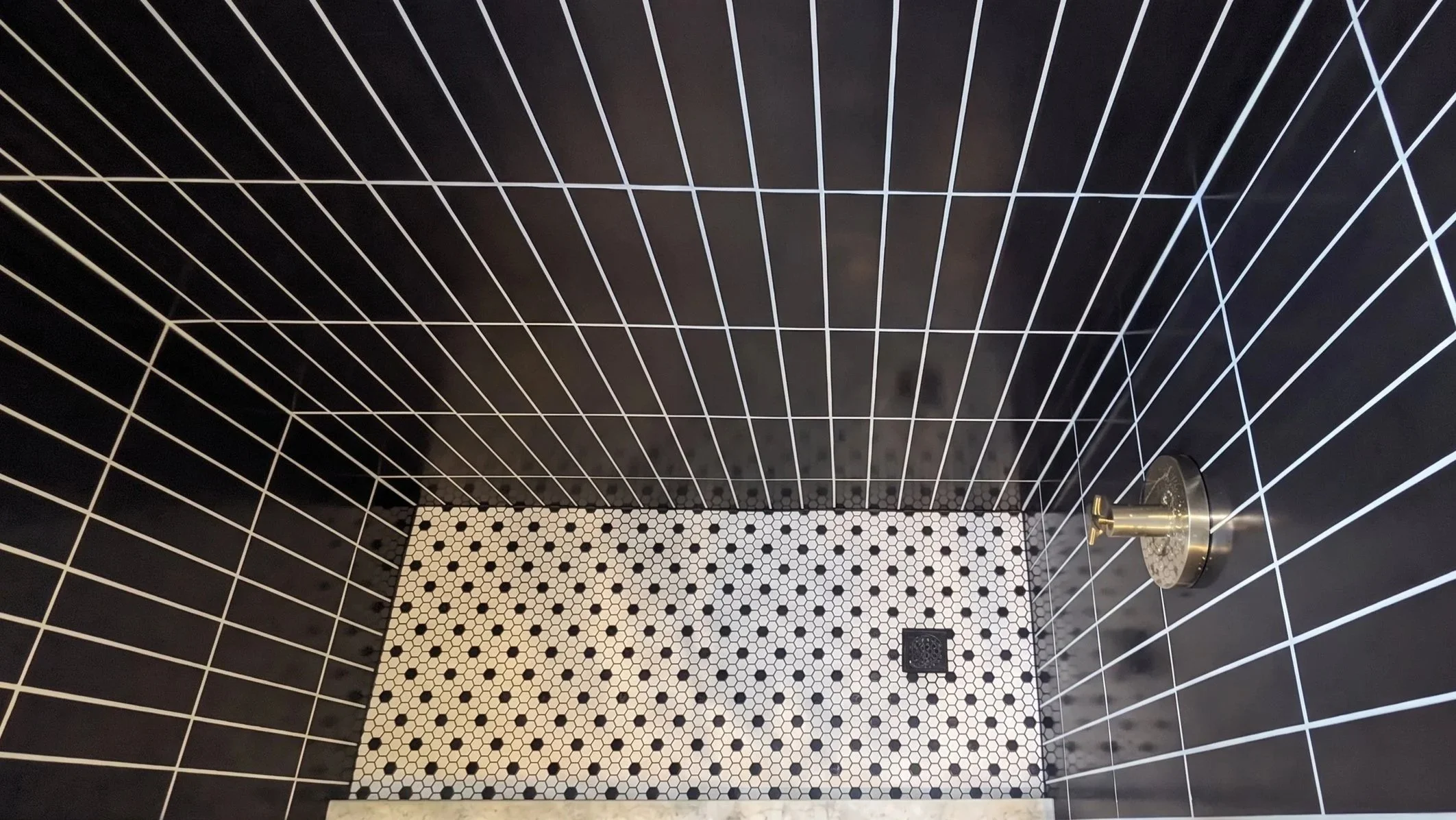 A black tiled shower with white grout lines, a circular chrome showerhead on the right wall, and a black and white patterned floor.