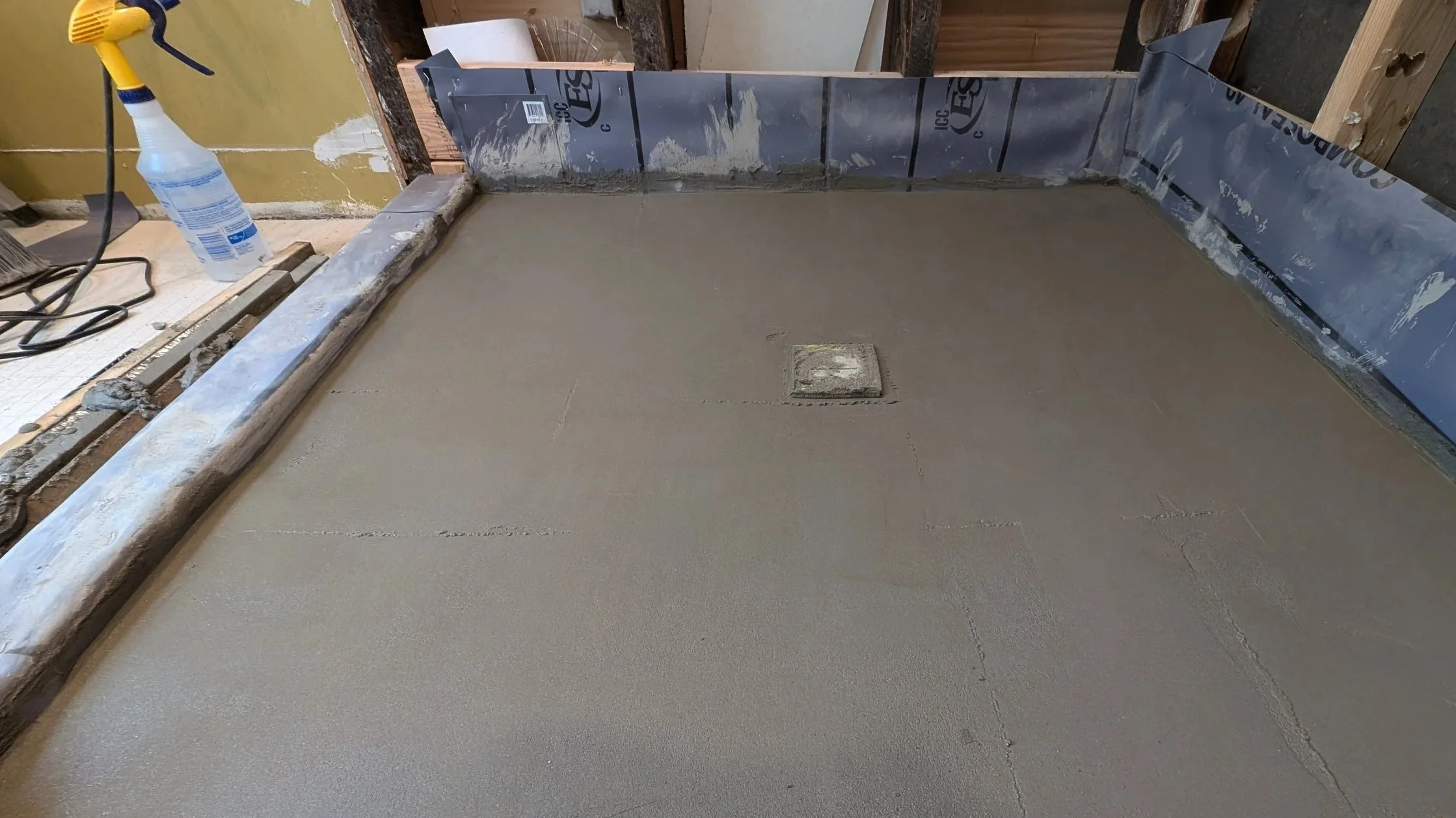 A construction site with a freshly poured concrete floor, an exposed pipe in the center, a plastic spray bottle, and construction tools and materials around the area.