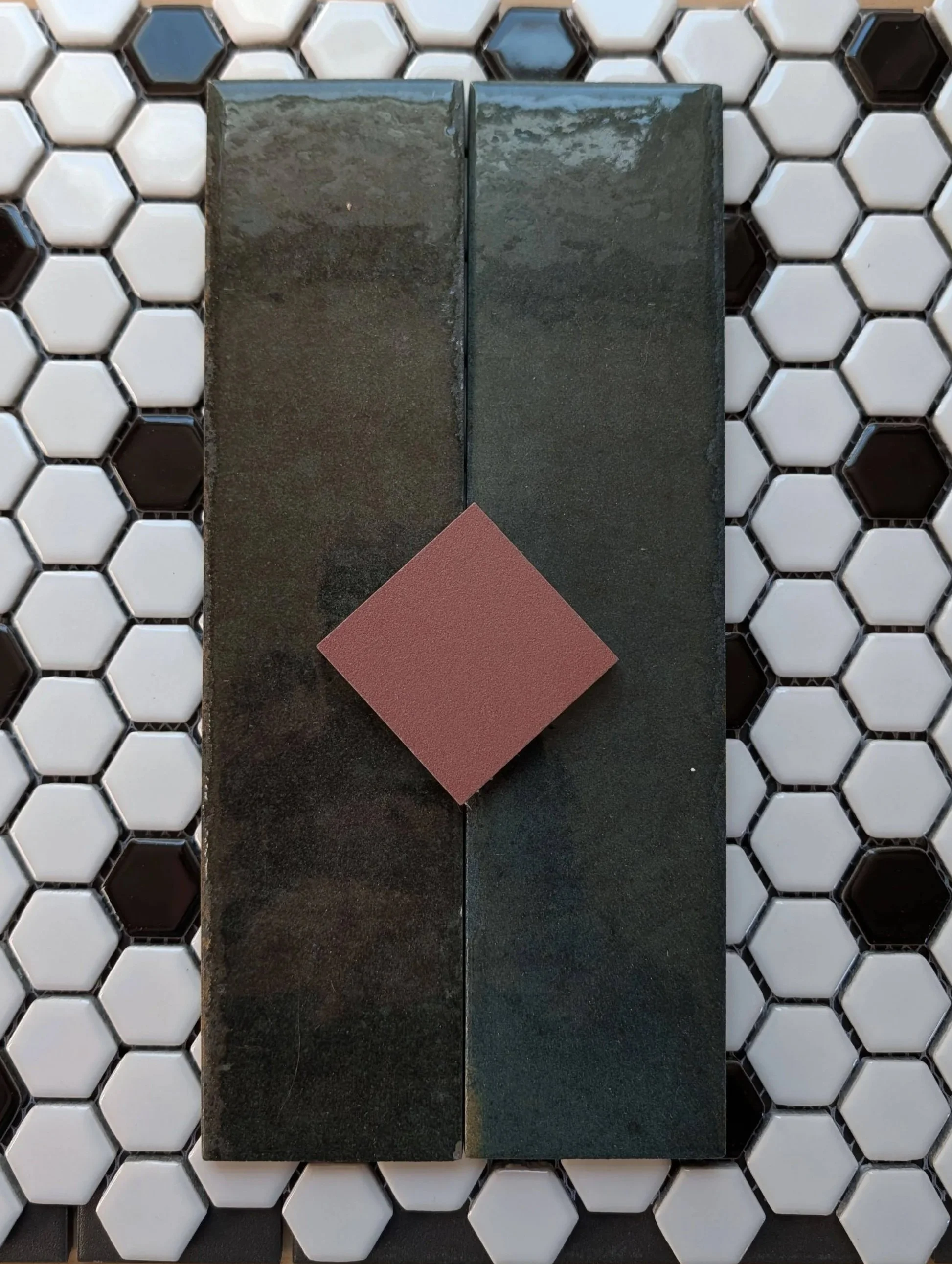 Two rectangular dark tile samples placed side by side on a black surface, with a smaller pink tile sample in front of them, all on a white hexagonal tile background.