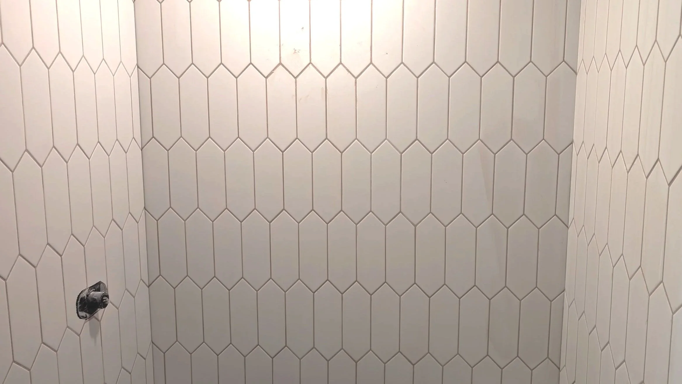 White hexagonal tile wall with a partially installed black and chrome shower valve and plumbing fixtures.