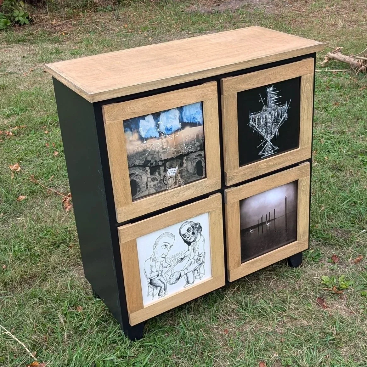 A black wooden cabinet with four drawers, each featuring a unique design or image framed with light wood. The cabinet is outdoors on grass with some greenery and twigs nearby.