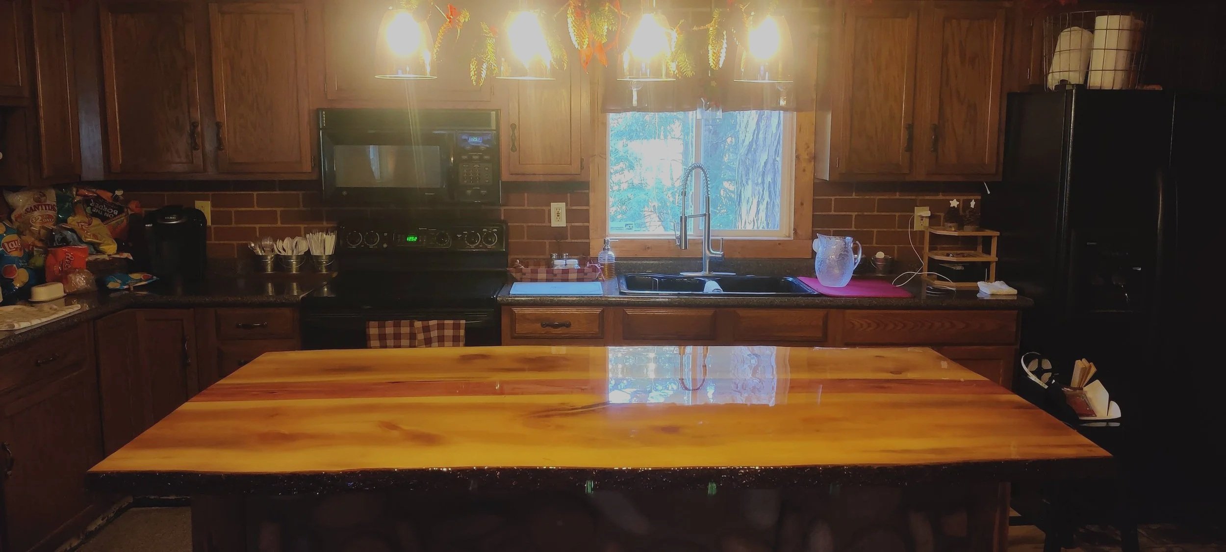 A kitchen with wooden cabinets, black appliances including a microwave and refrigerator, a window above the sink, and a large wooden table in the foreground.