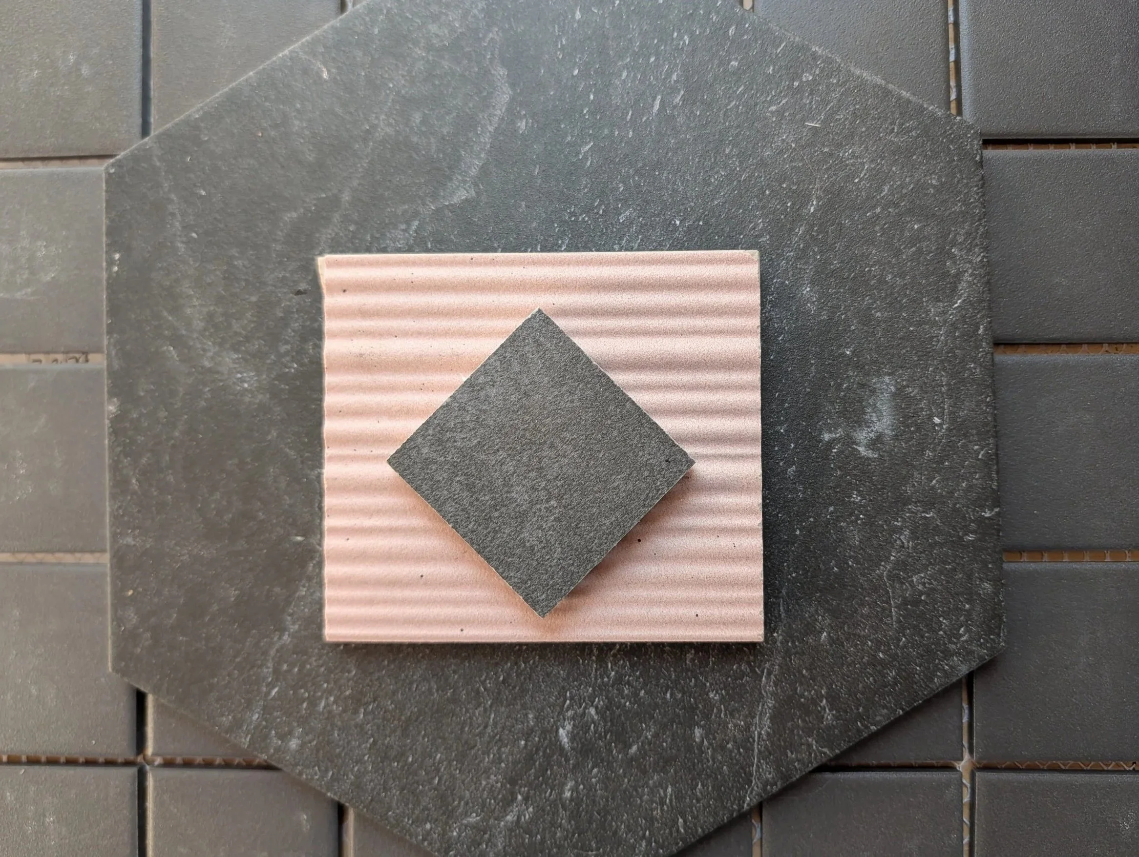 Close-up of various textured tiles and stone pieces arranged on a surface, including dark gray, pink, and darker gray materials.