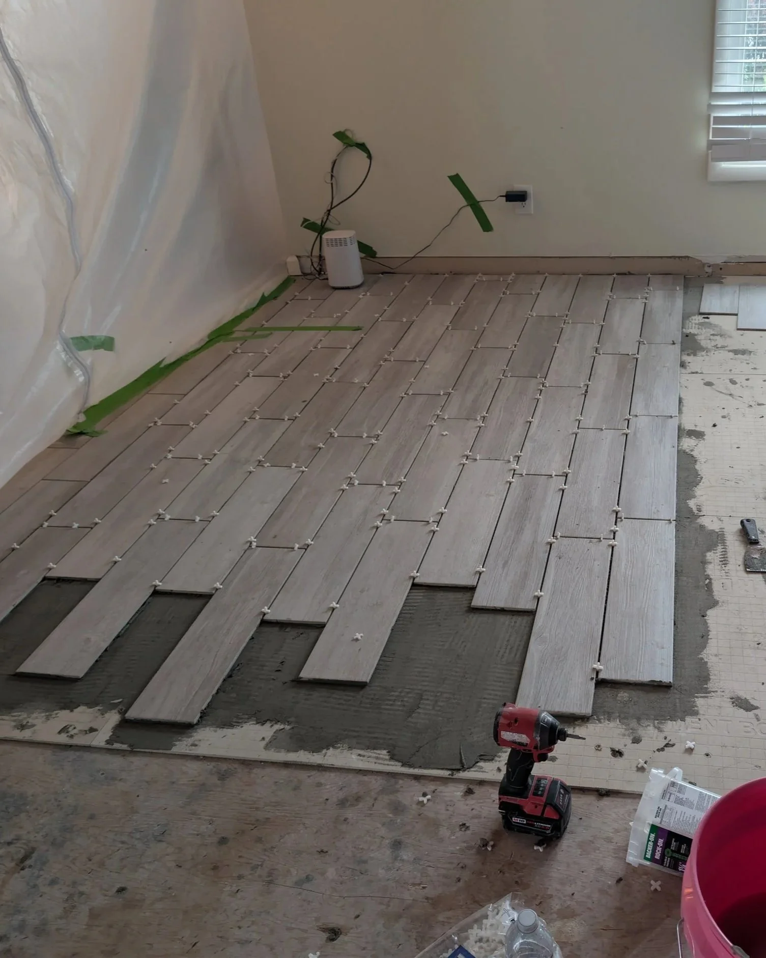 Room with flooring installation in progress, showing wooden planks being laid with spacers, various tools, a drill, and a water bottle on the unfinished floor.