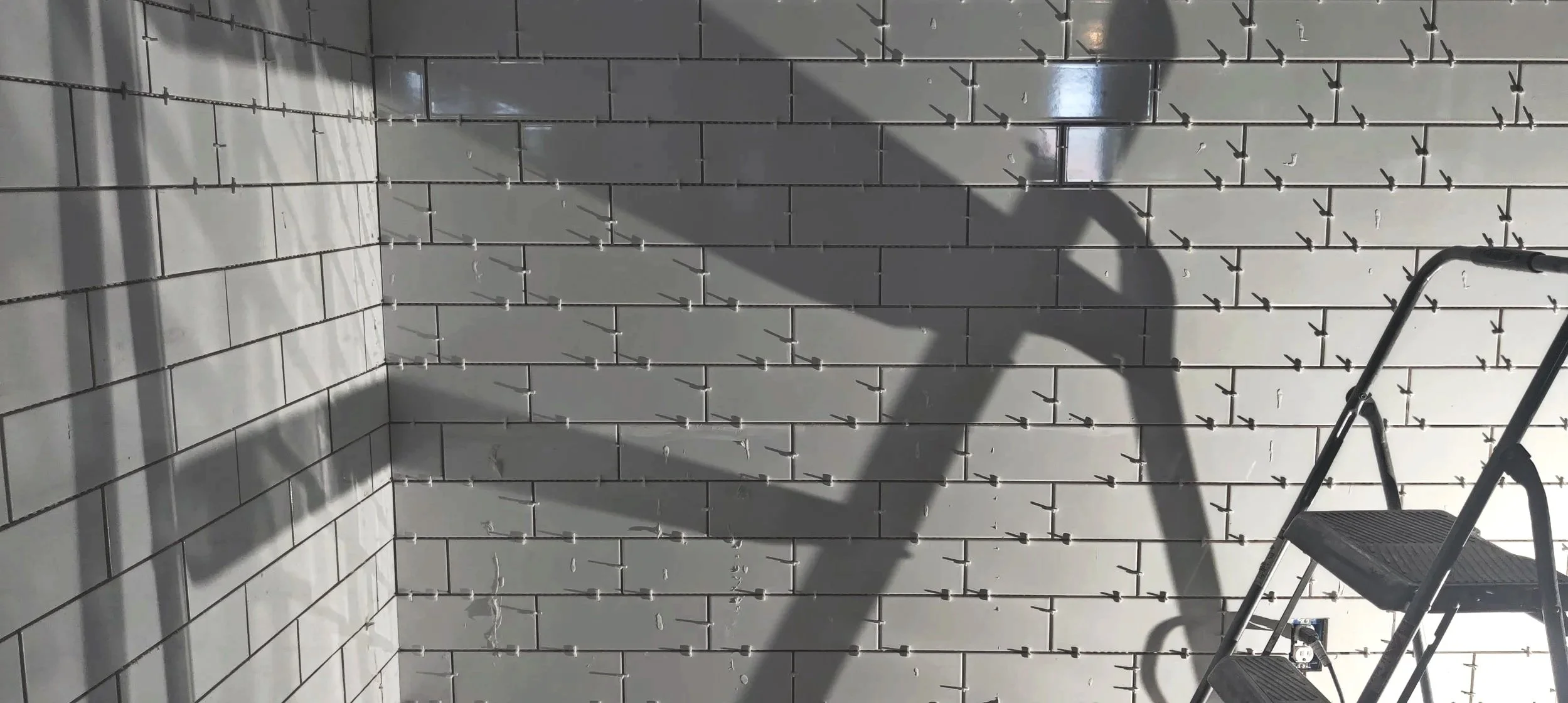 A wall with metal wall anchors protruding out, arranged in a grid pattern. There is a stepladder on the right side of the image with shadows cast on the wall.