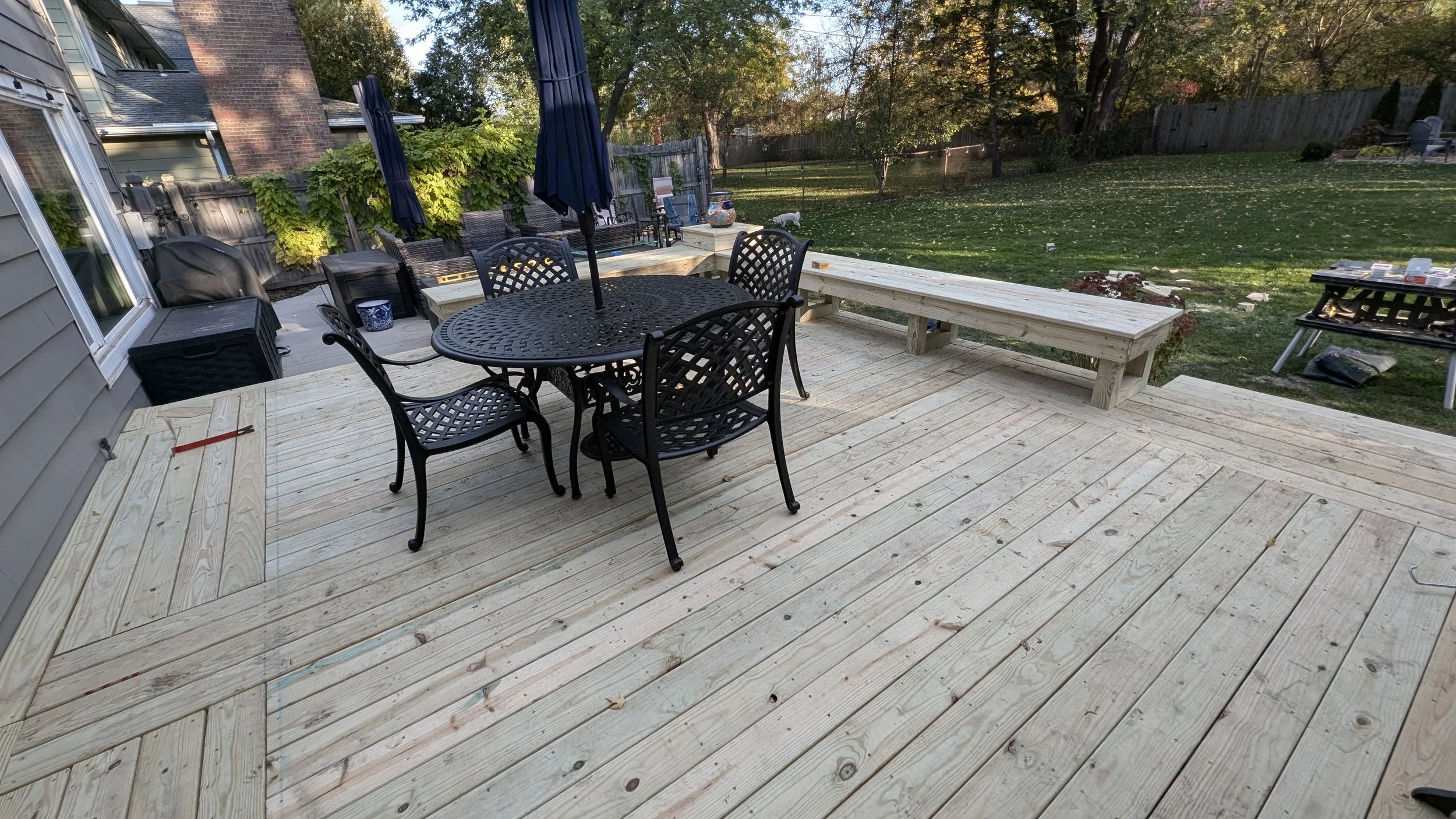 Backyard wooden deck with patio furniture, including a round table with chairs and a bench. The yard has grass, trees, and a fence in the background, with some outdoor items like tomato cages and a grill.