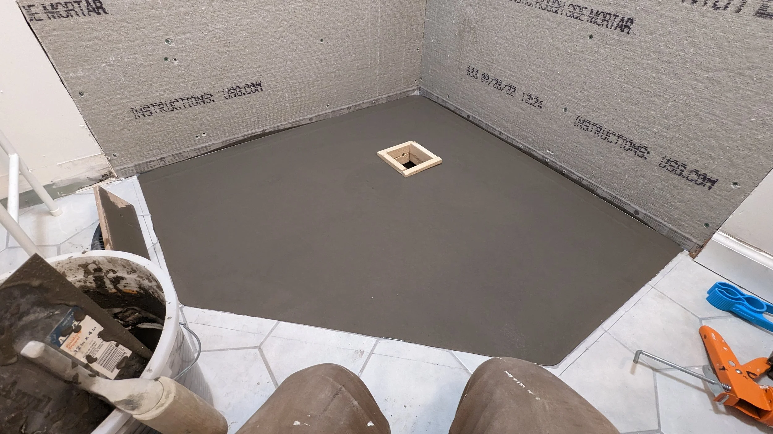Bathroom floor with newly poured concrete, surrounded by construction tools and materials, including pipes and a level, with unfinished drywall on the walls.