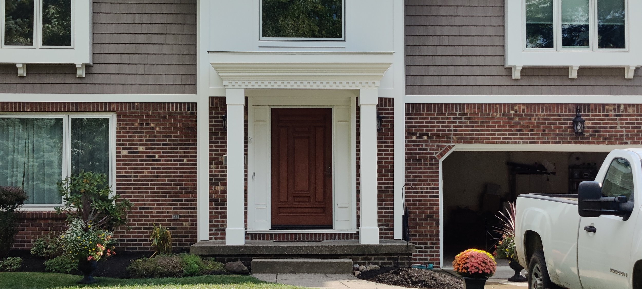 Front entrance of a house with a wooden door framed by white columns, brick and siding exterior, large windows, a small porch with steps, a white truck parked nearby, and flower pots with colorful flowers in the yard.