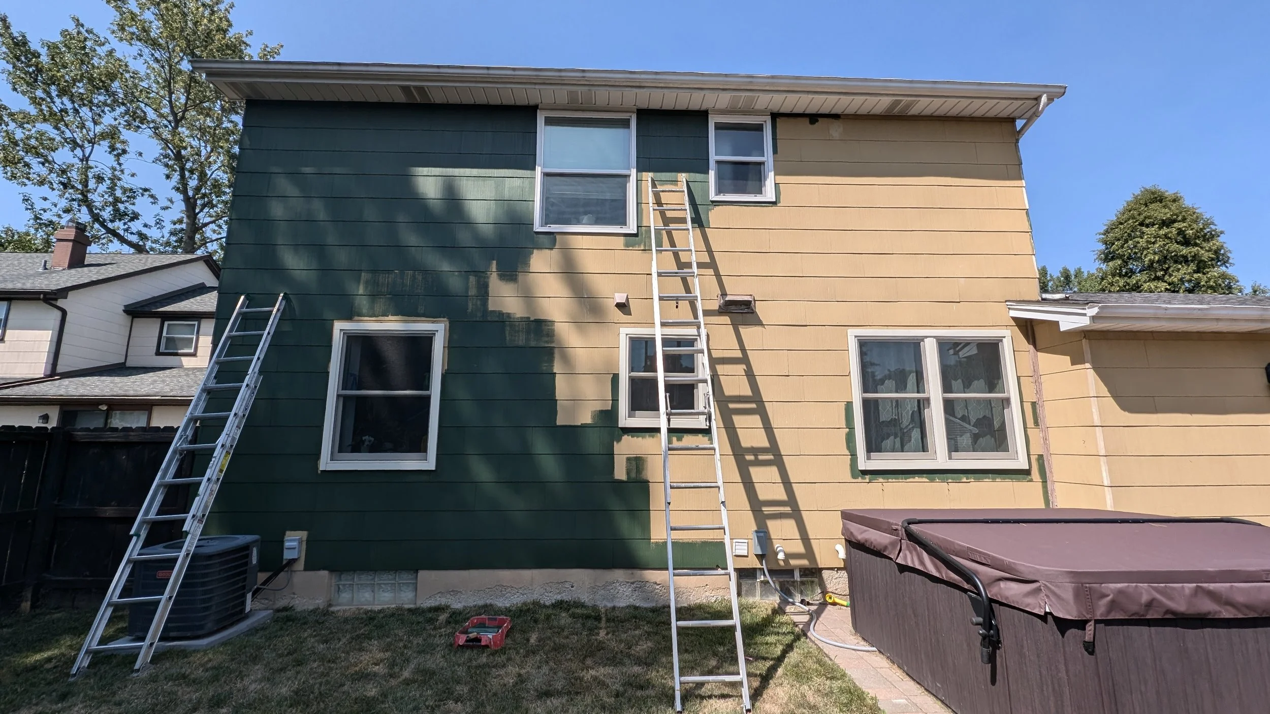 Two ladders leaning against the side of a house with green and beige siding, as part of a house renovation or painting project.
