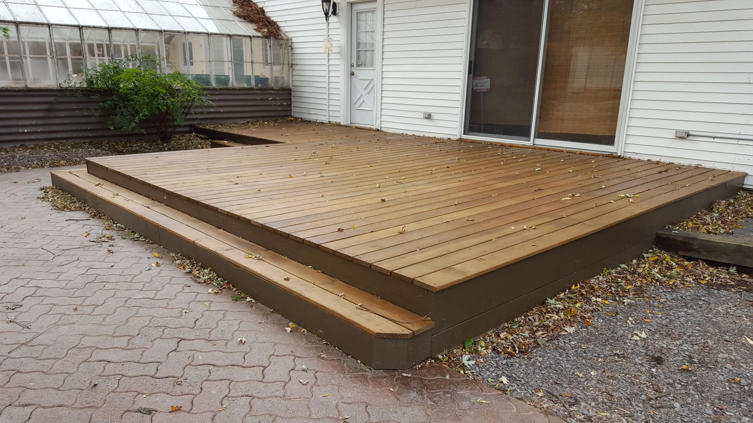 Wooden deck outside a house with white siding, a sliding glass door, and a white door, surrounded by fallen leaves and a small bush.