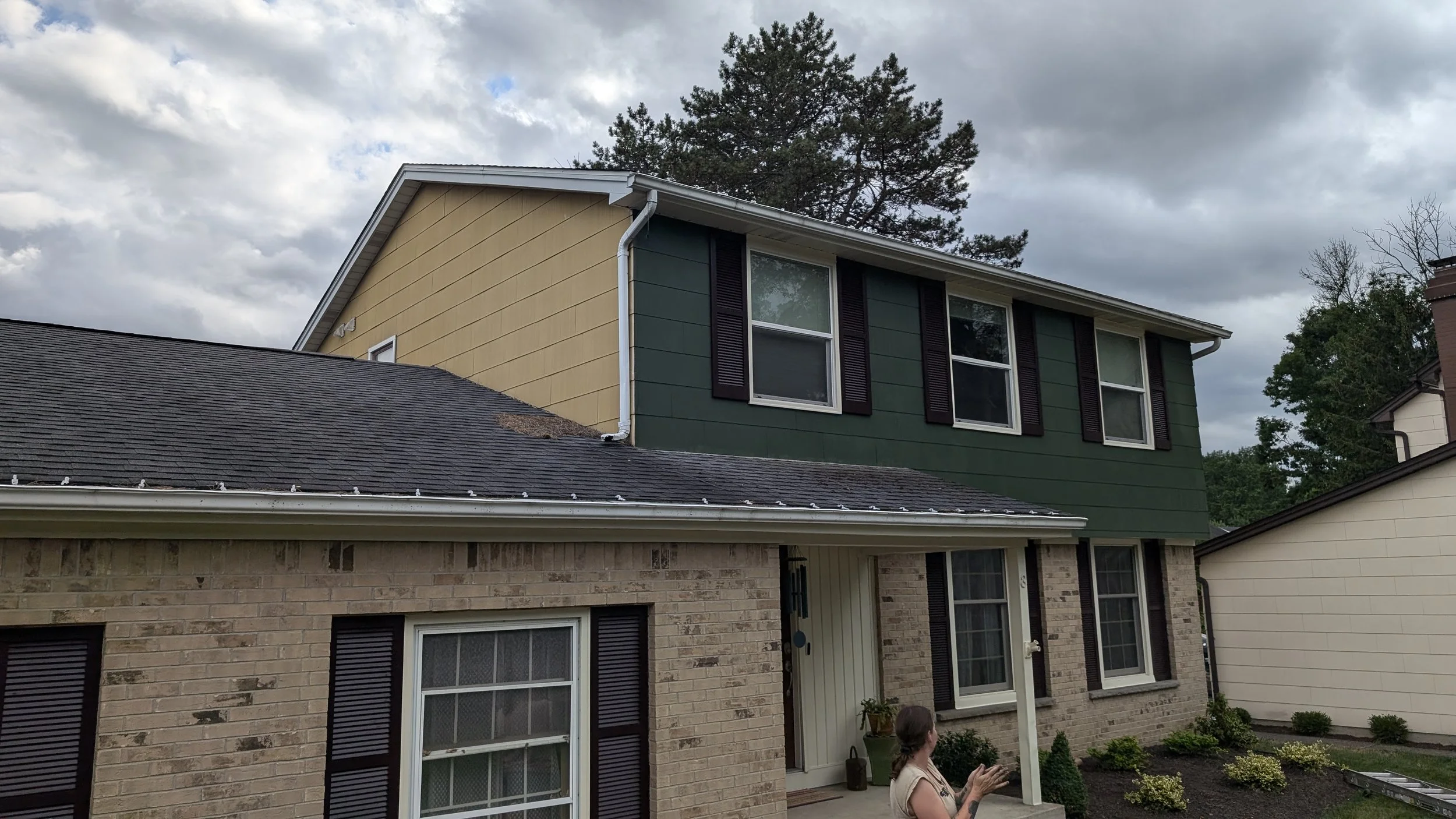 A two-story house with a brick lower level and green siding on the upper level. The house has several windows with dark shutters, and a woman stands outside near a sliding glass door. The roof is dark gray, and there are trees and a cloudy sky in the