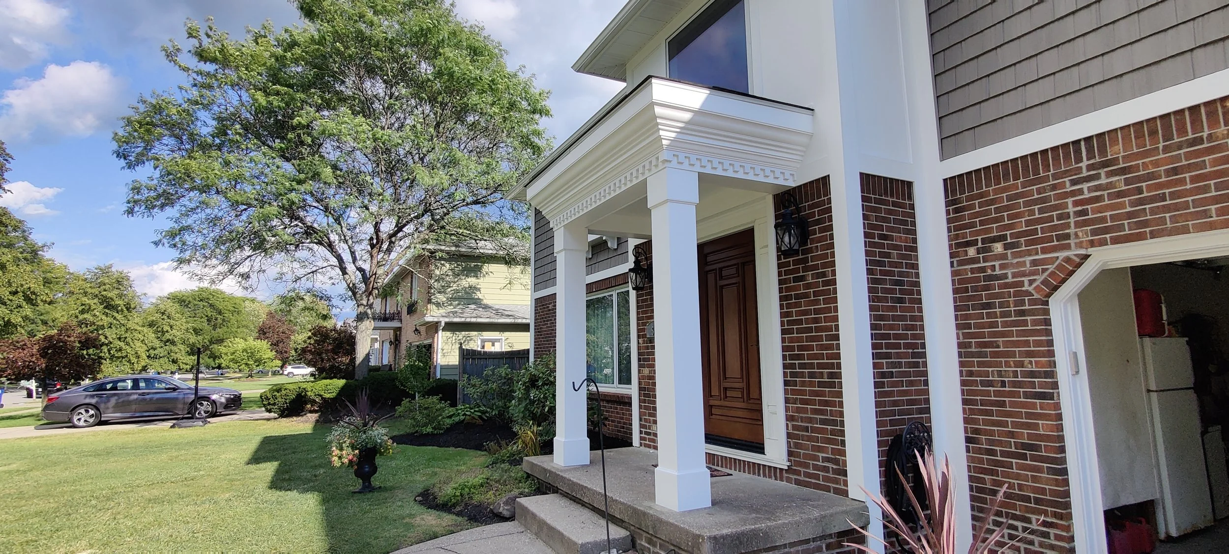 Front porch of a brick house with columns, a wooden door, and outdoor lanterns, with a lawn and trees in the background.