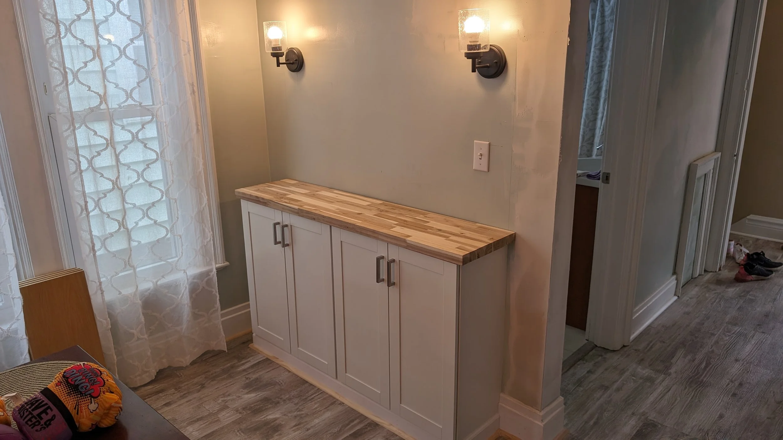 A beige hallway with a white cabinet with a wooden top beneath the wall-mounted lights. A window with lace curtains is on the left, and there is a door on the right with shoes visible on the floor.