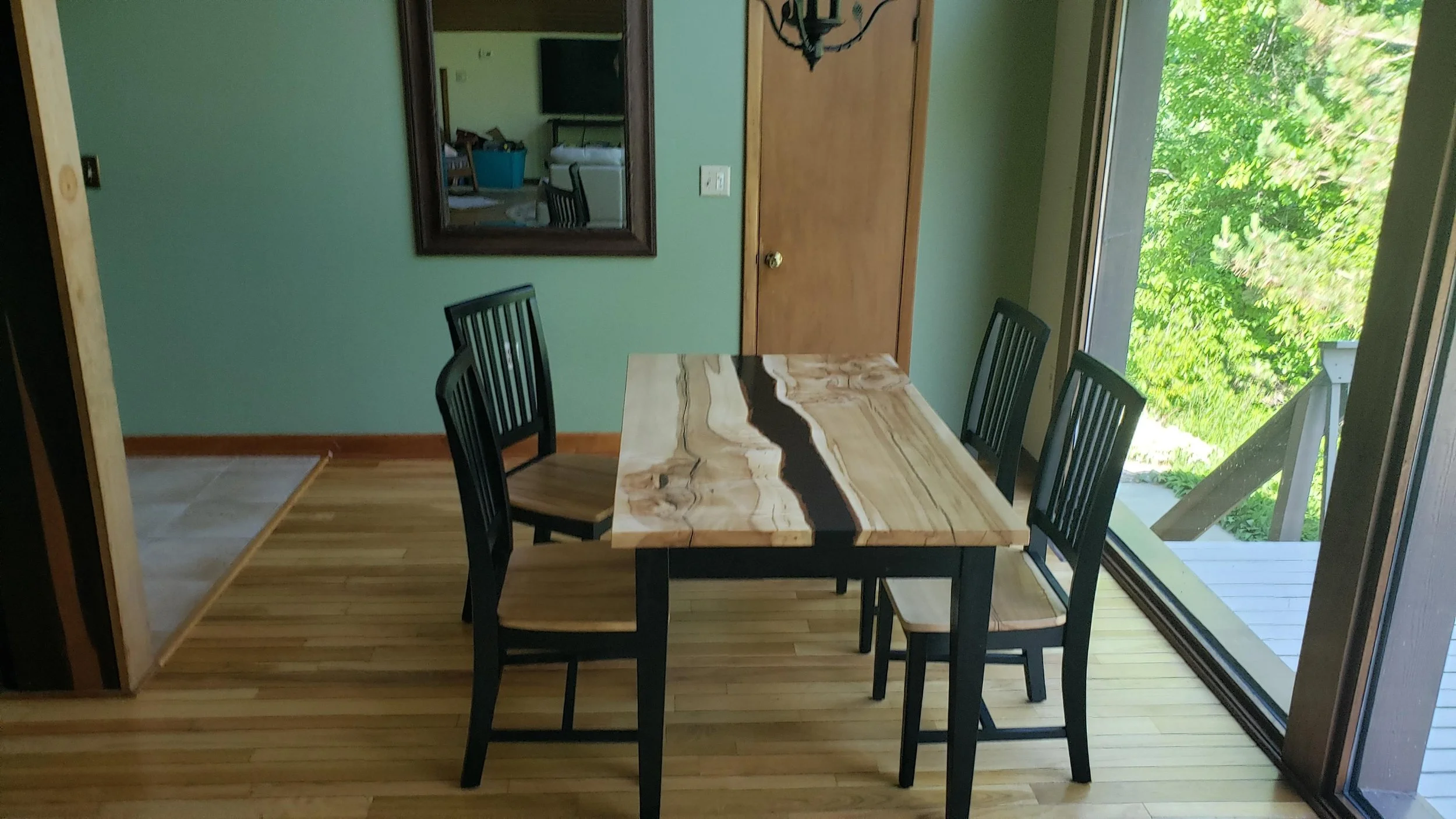 Dining area with a wooden table featuring a natural edge, surrounded by five black chairs, next to a large sliding glass door showing green trees outside. A mirror and a closed wooden door are visible on the green-painted wall.