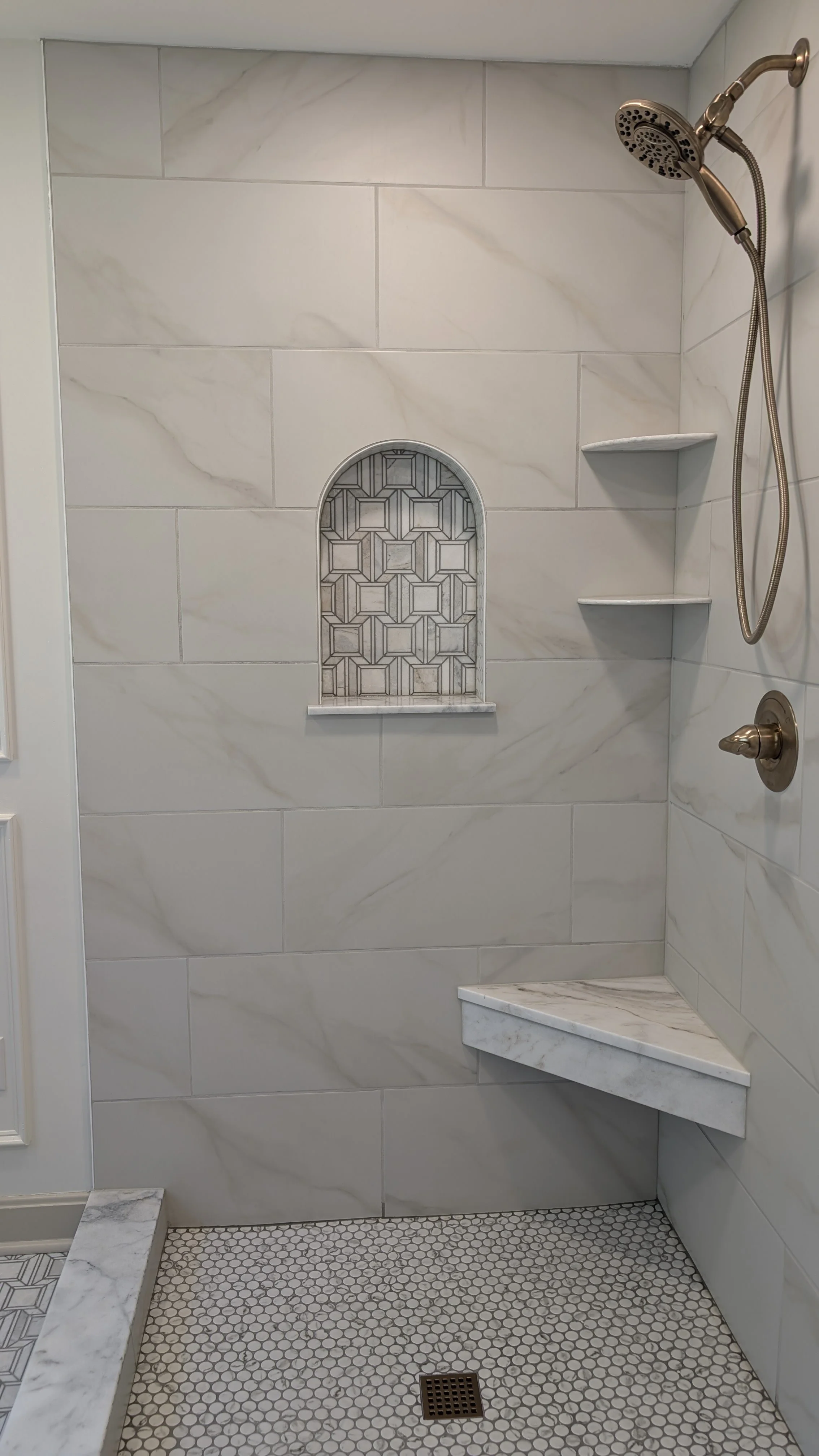A tiled shower with large marble-like wall tiles, a built-in shelf with patterned tile, small hexagon floor tiles, a corner marble bench, and a bronze showerhead and control knob.