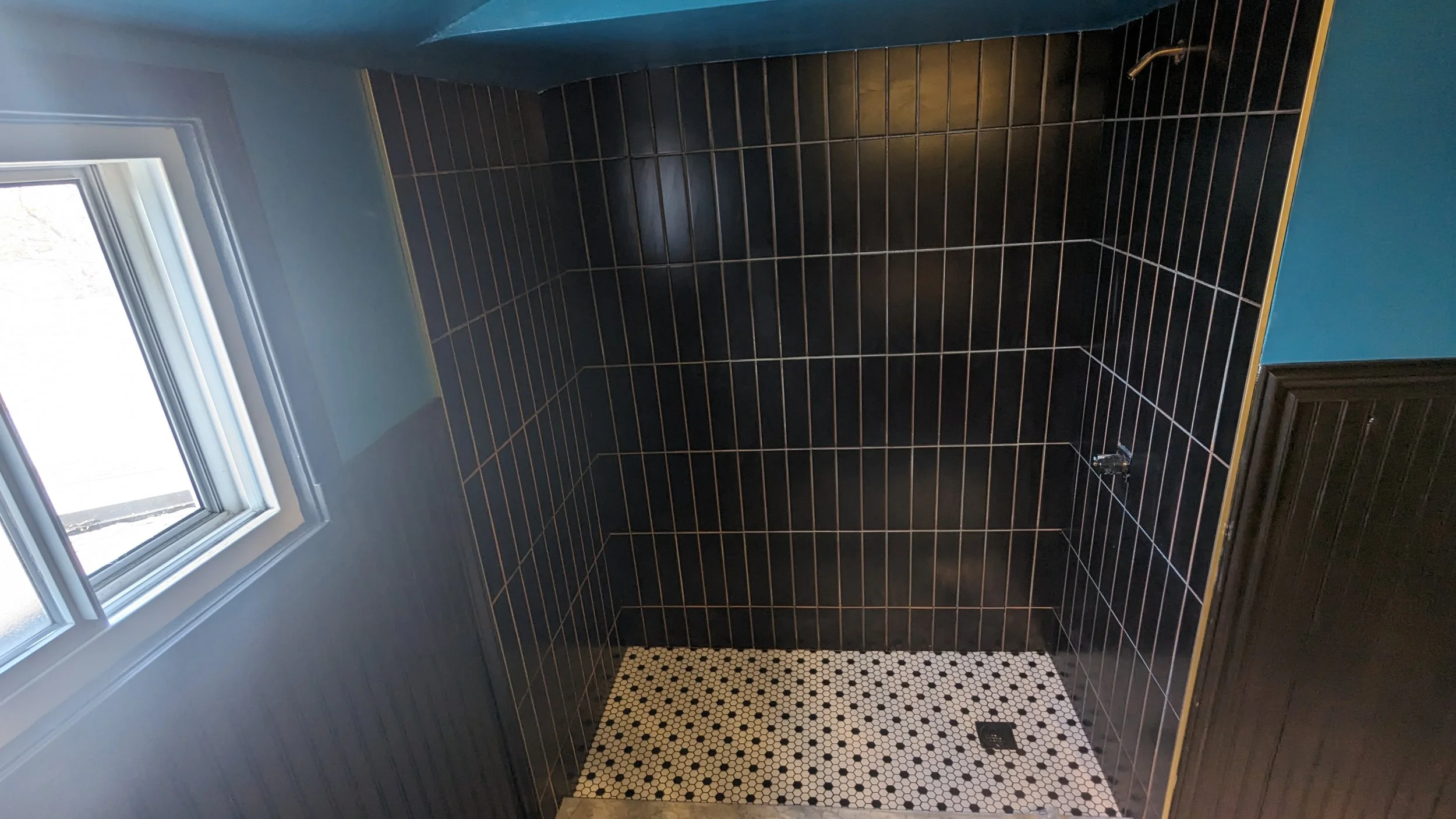 Empty shower with black tiles, a small window, and black and white hexagonal floor tiles.