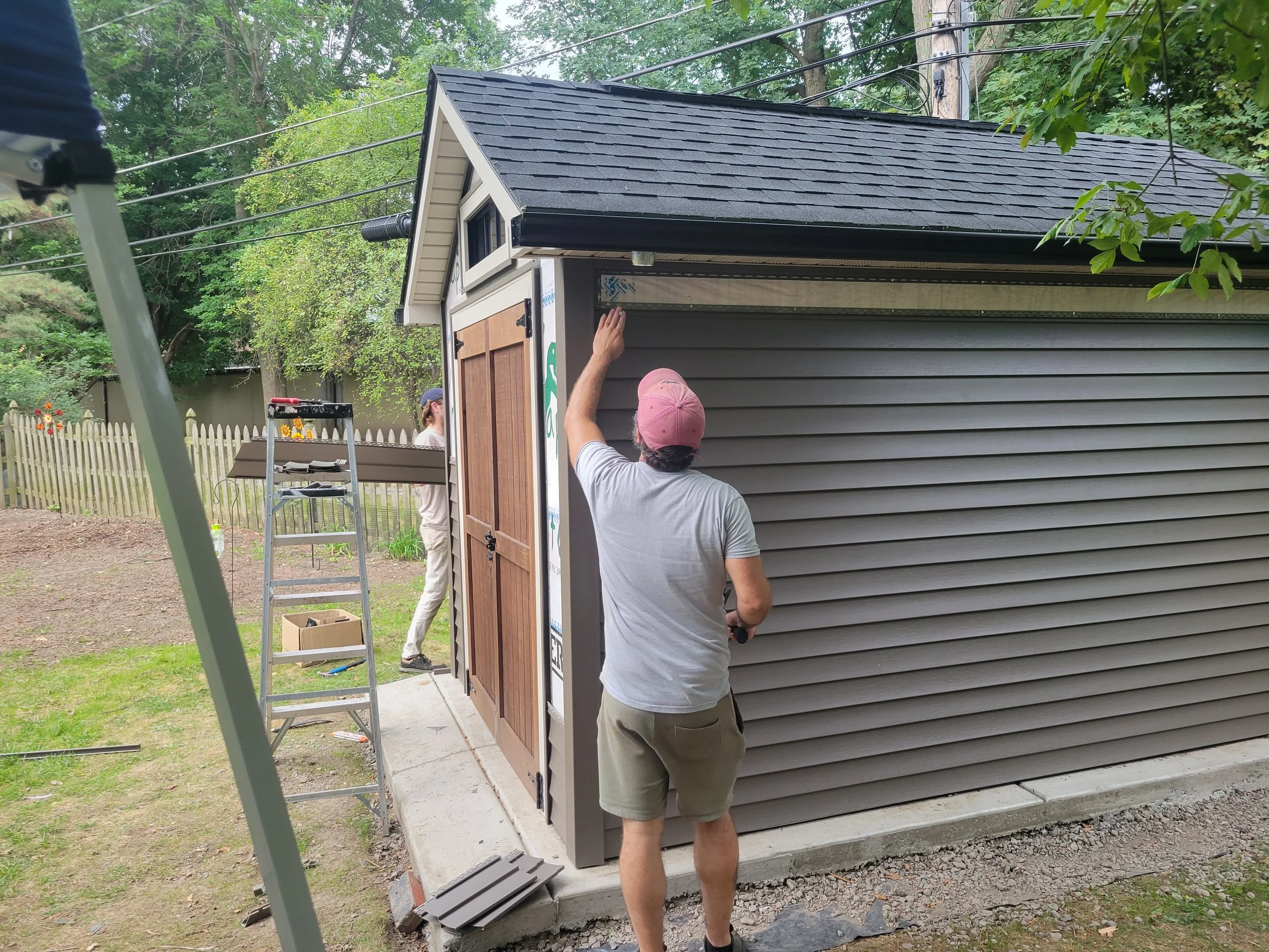 People installing siding on a small building with a gabled roof, adjacent to a wooden fence and surrounded by green trees.