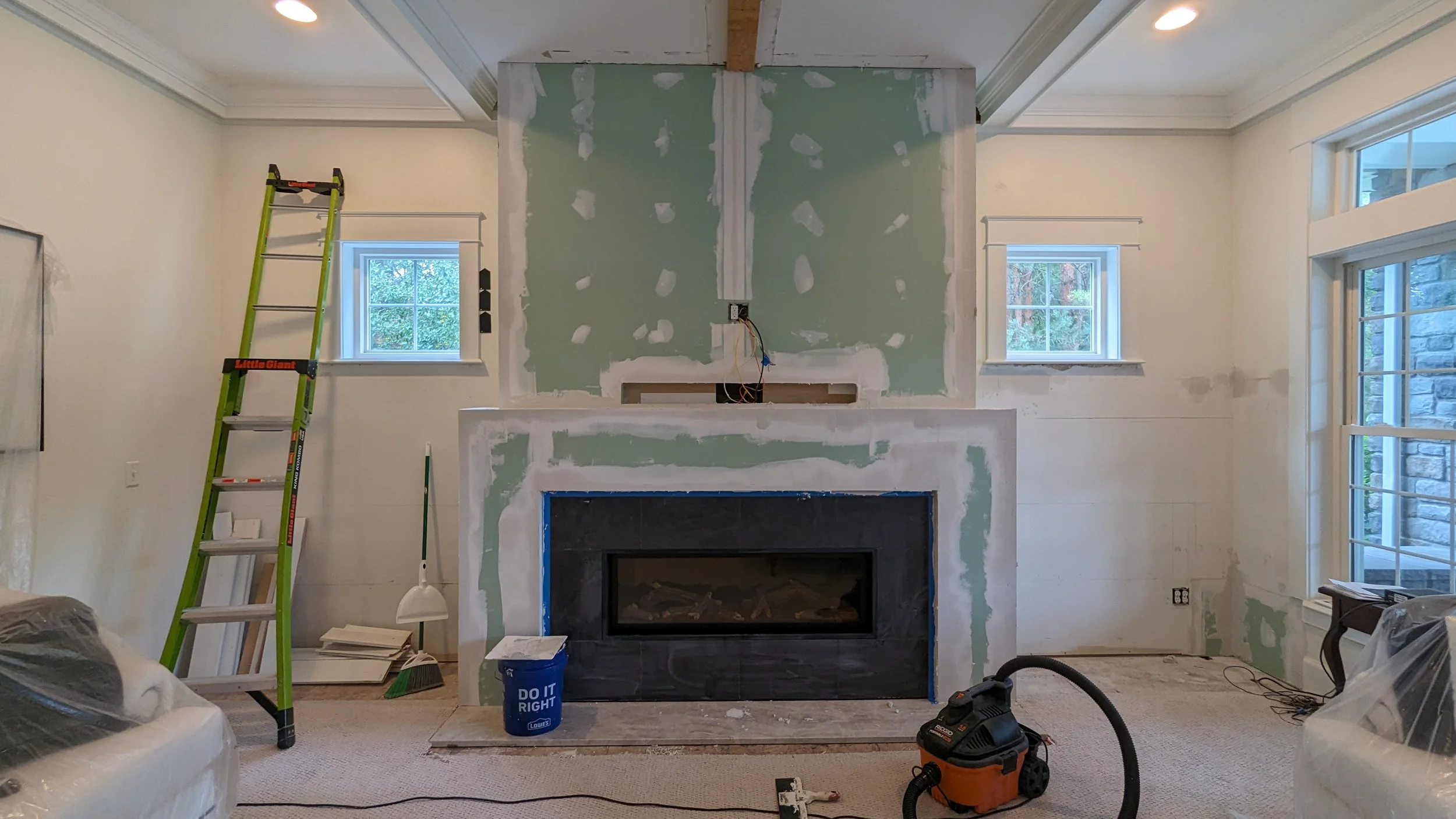 Living room under renovation with a fireplace, partially finished green drywall around the fireplace, a yellow ladder on the left, construction tools including a shop vacuum, buckets, and a pile of papers, and two windows on either side of the firepl