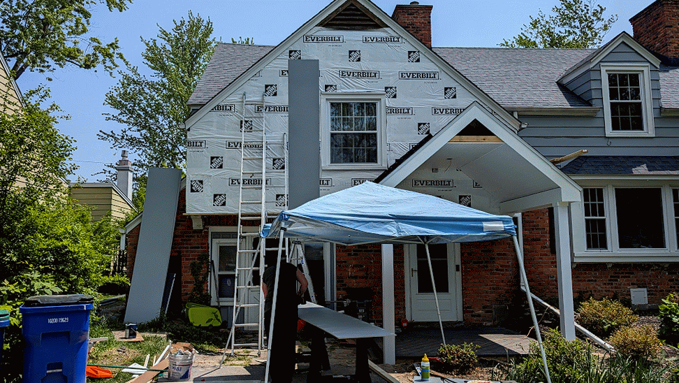 A house under renovation with a portion of the exterior siding removed, workers present, and construction equipment set up outside.