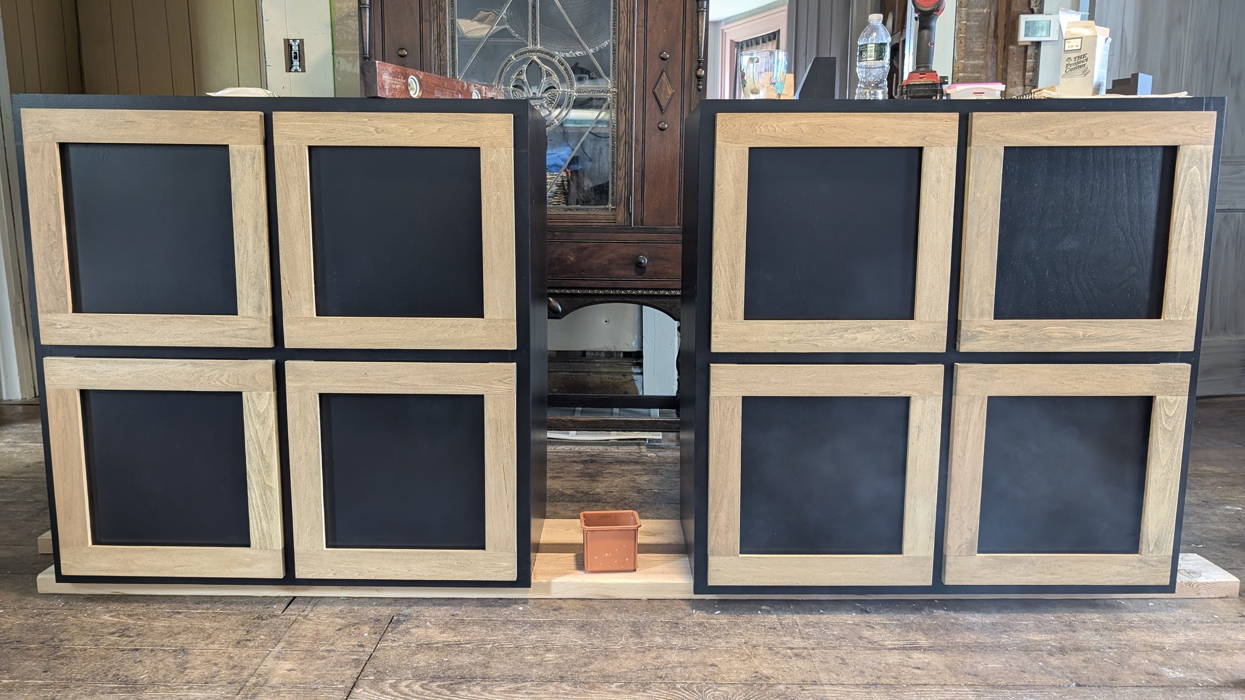 Two unfinished wooden and black framed cabinets or furniture pieces in a workshop, with a small orange container on the floor and various workshop tools and items on the countertop in the background.