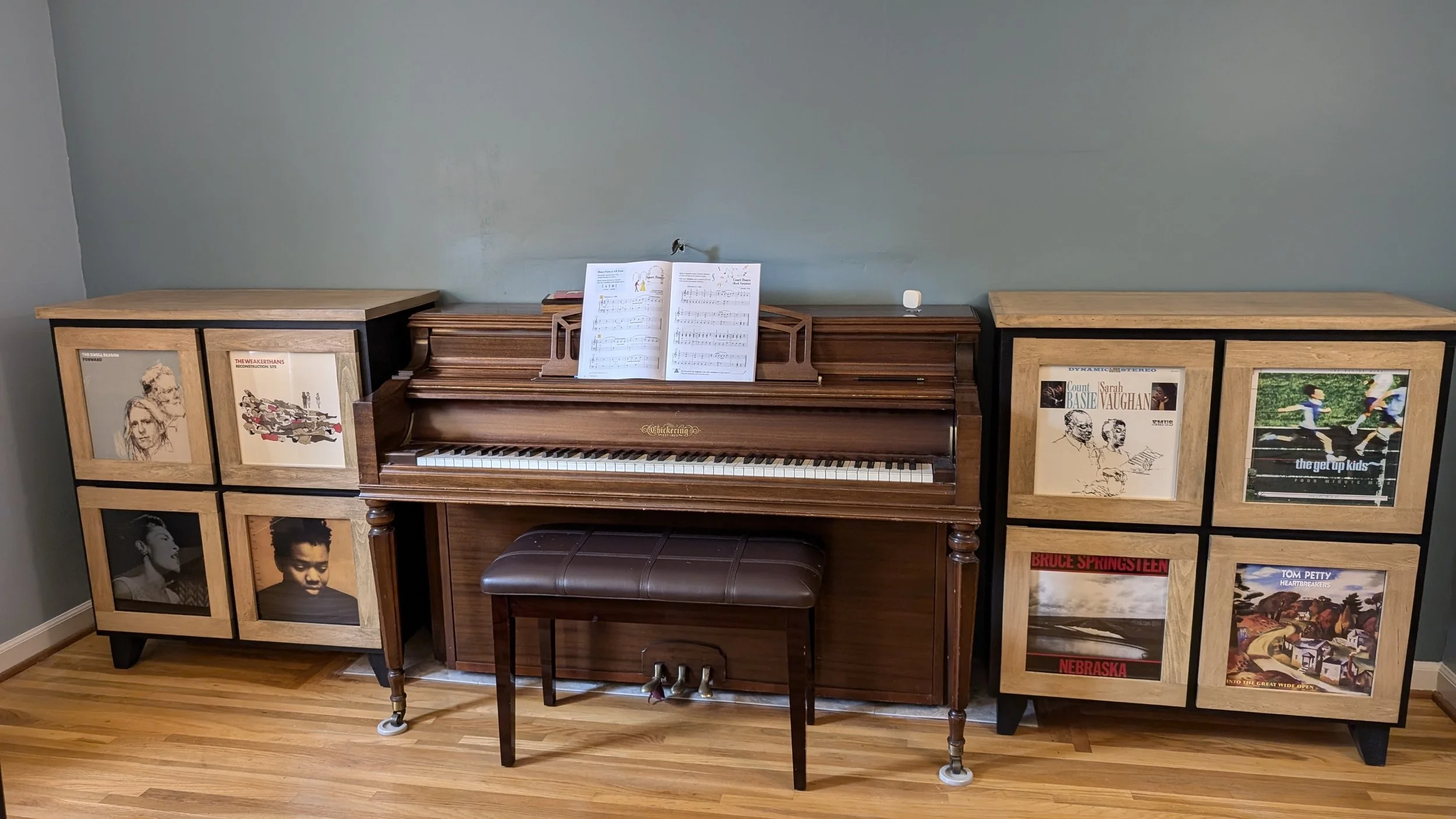 A wooden piano with open sheet music on top, flanked by two cabinets decorated with album covers and photographs, set against a light grey wall and wooden floor.
