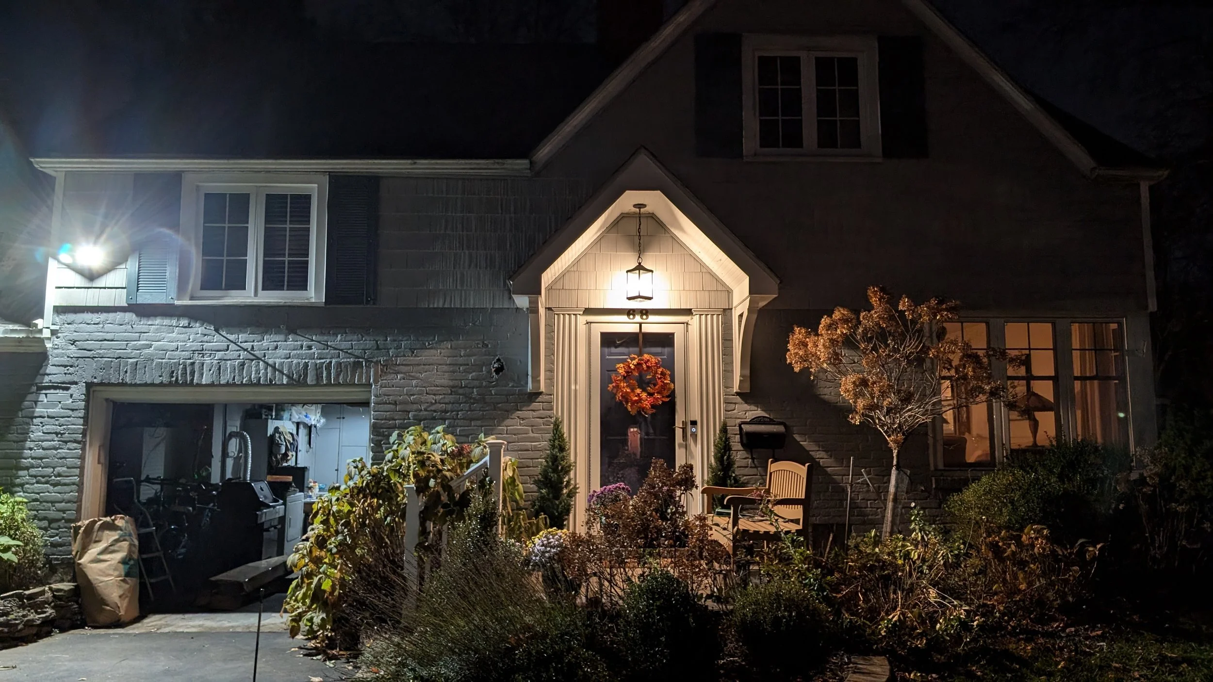 A two-story house at night illuminated by exterior and interior lights. The house has brick and siding exterior, with a front porch decorated with a fall wreath on the door. Landscaping includes small trees and bushes, and there are items like a gril