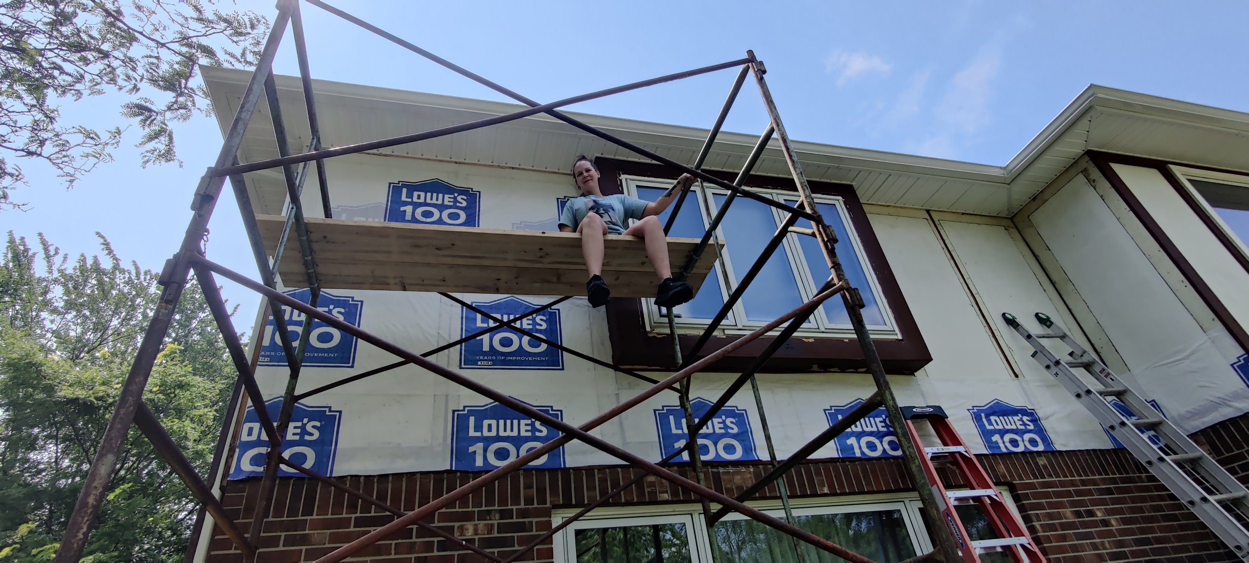 A person sitting on a wooden platform in the middle of a construction scaffold outside a house under renovation, with Lowe's 100 years of improvement signs visible.