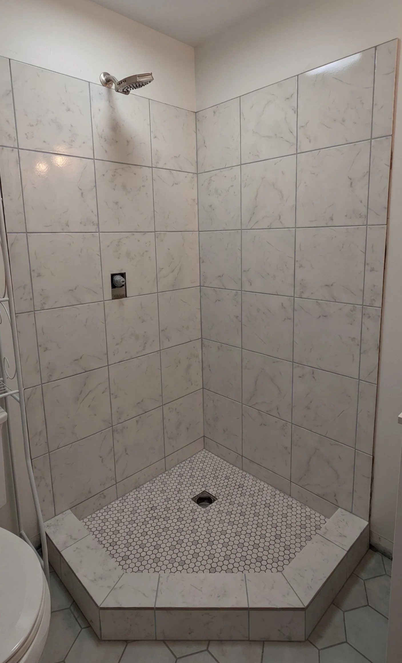 Bathroom shower with white marble tiles, a handheld shower head, a drain on the tiled floor, and a small step with a marble edge.