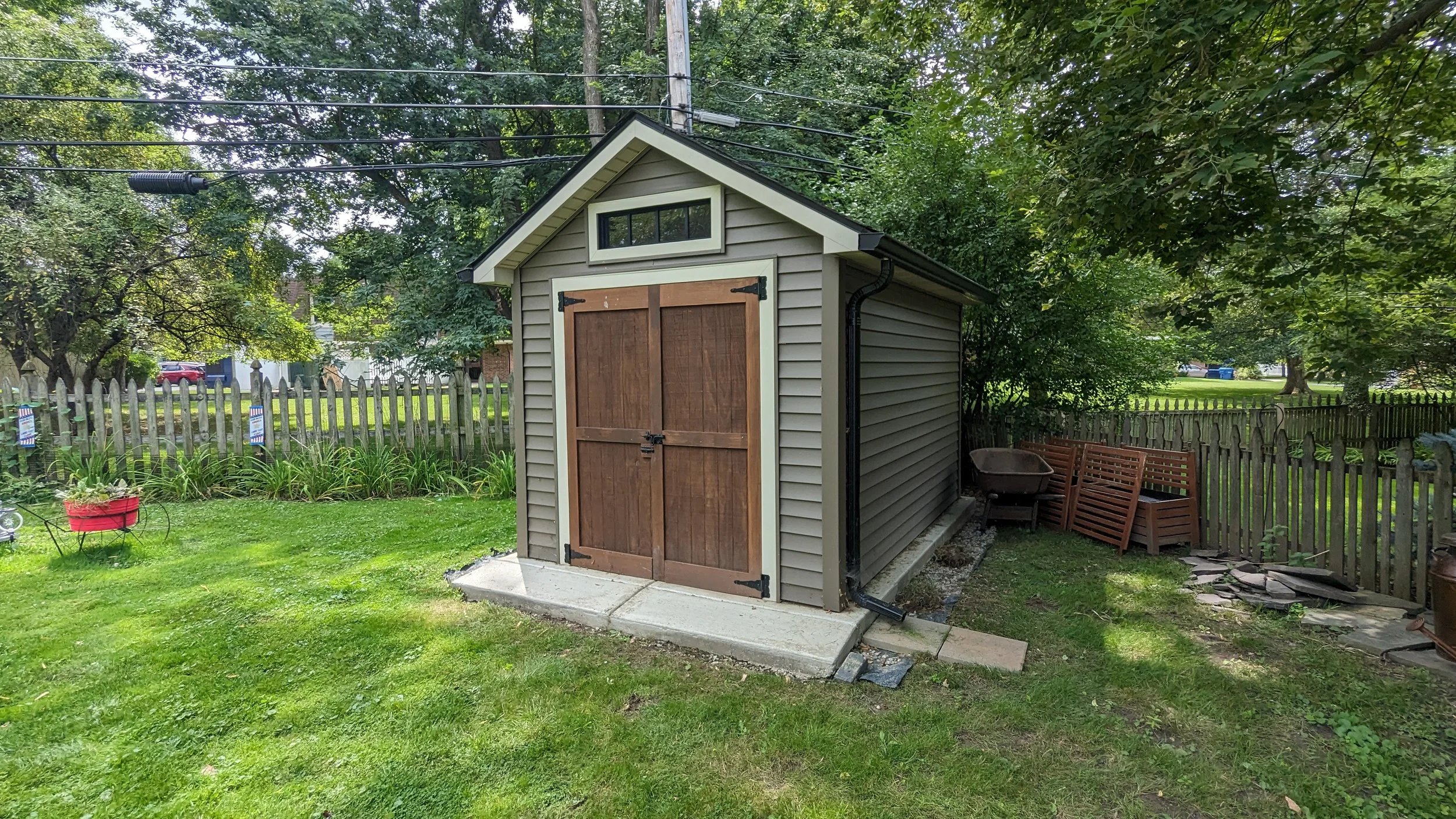 A small backyard shed with double wooden doors, gray vinyl siding, and a black roof, surrounded by a green lawn and a wooden fence, with trees and outdoor furniture in the background.
