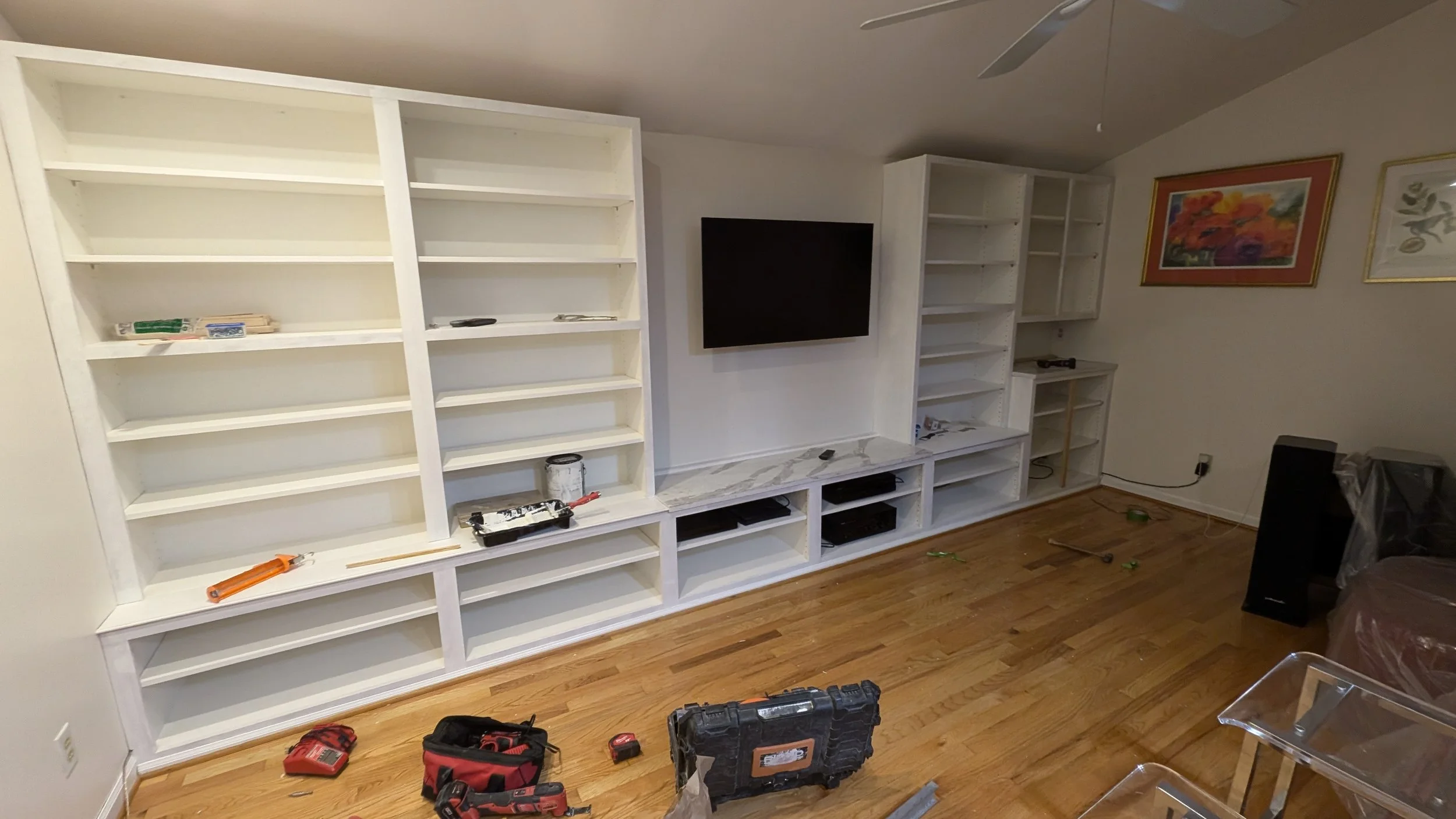 Room with white built-in bookshelves, television, pictures on the wall, and various tools and equipment for painting or renovation on the floor.