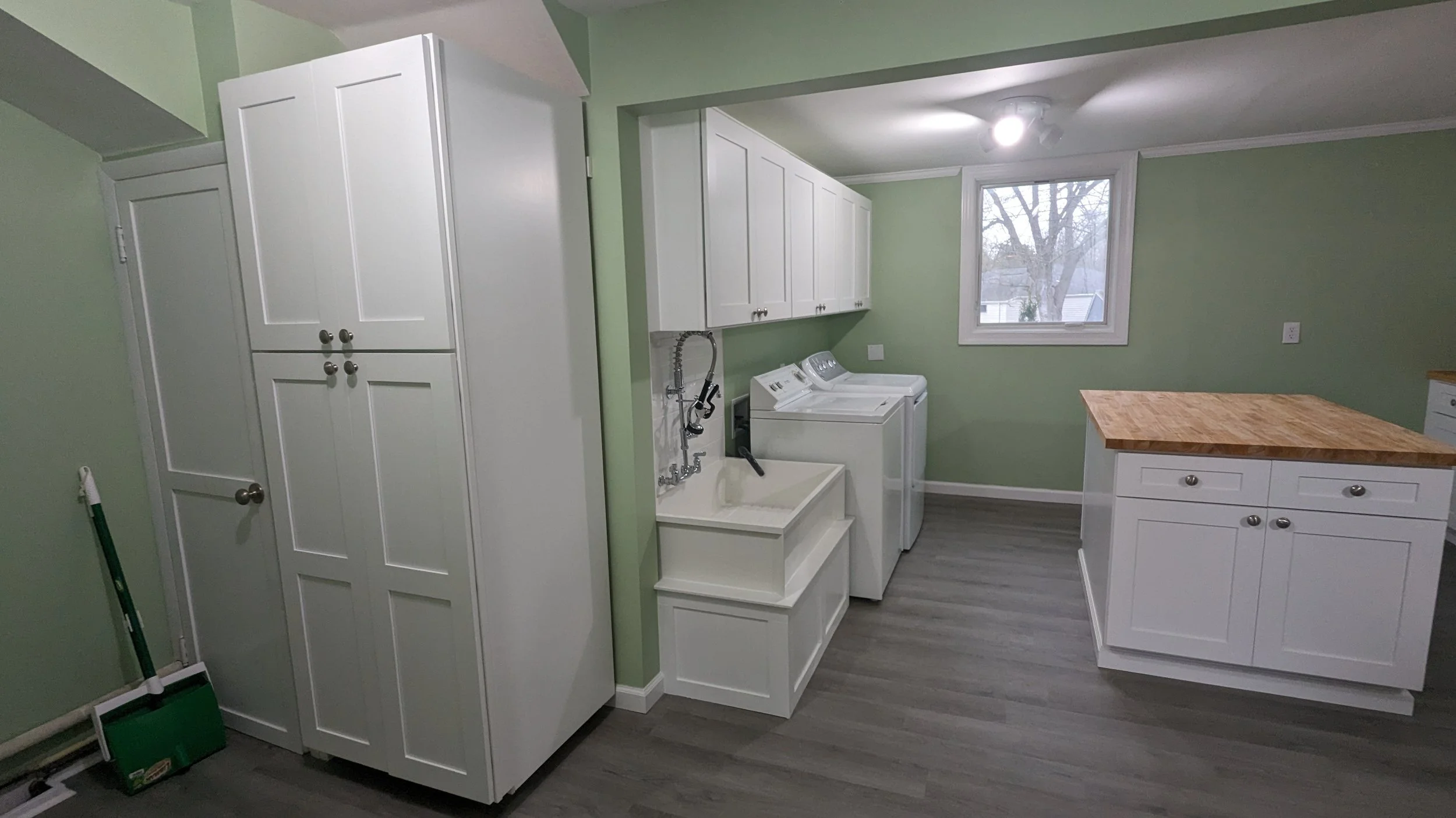 Laundry room with white cabinets, a deep sink, washing machine and dryer, a wooden island with drawers, a window showing outside trees, and green walls.