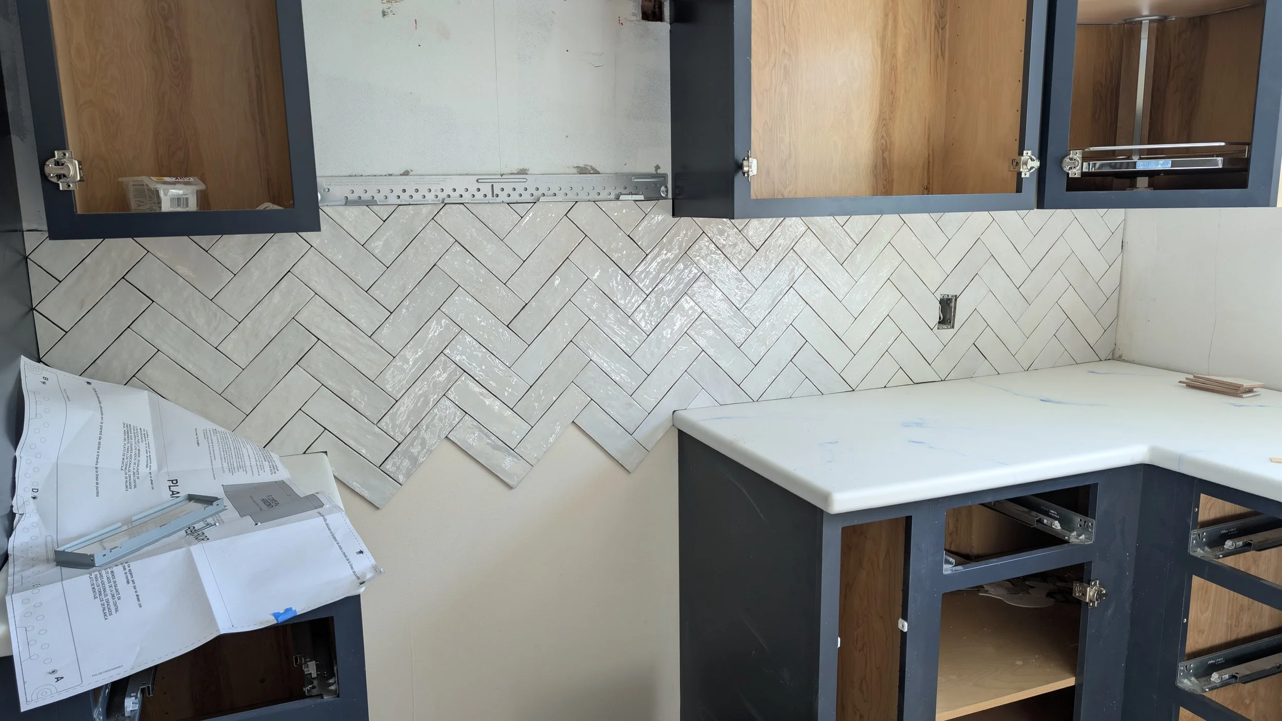 Cabinet frames with open spaces for drawers or doors, a partially installed herringbone tile wall, construction plans, and tools on a light-colored countertop in a kitchen renovation.