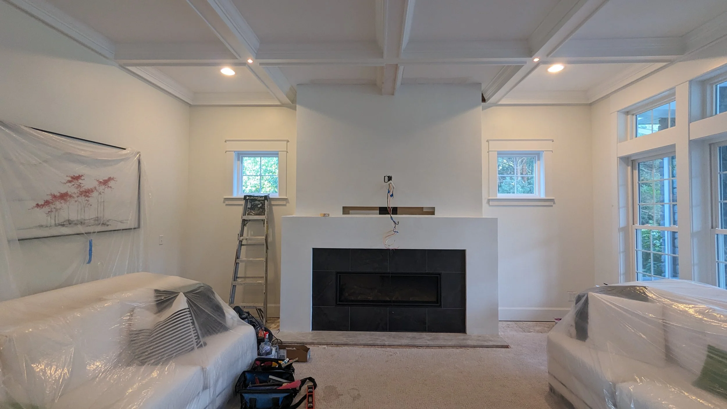 Living room under construction with white walls, a fireplace, multiple windows, and furniture covered in plastic. A ladder and tools are present, indicating ongoing work.