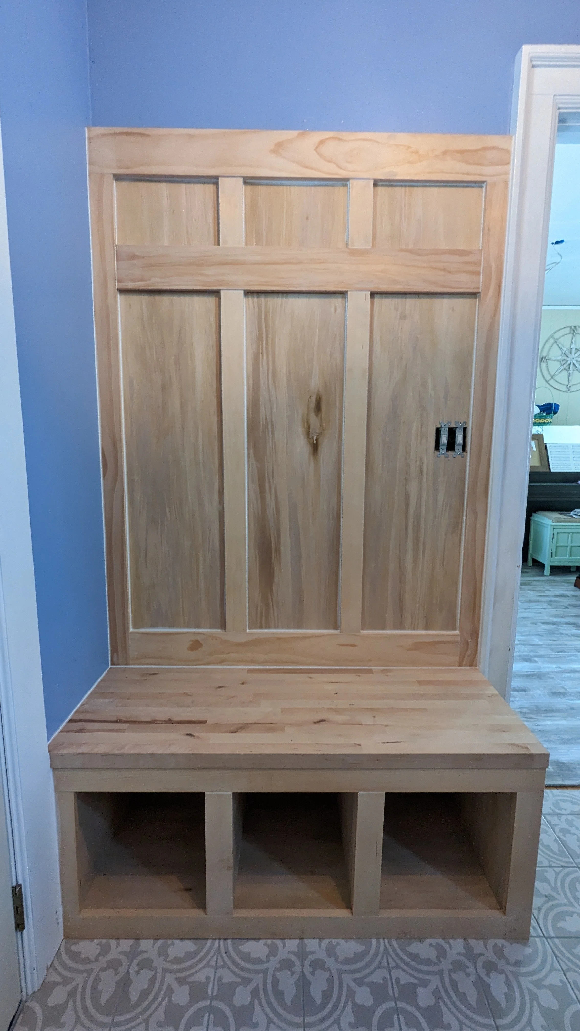 Wooden entryway with a bench, large panel door, and open cubby storage at the bottom, against a blue wall with a white door frame on the right.