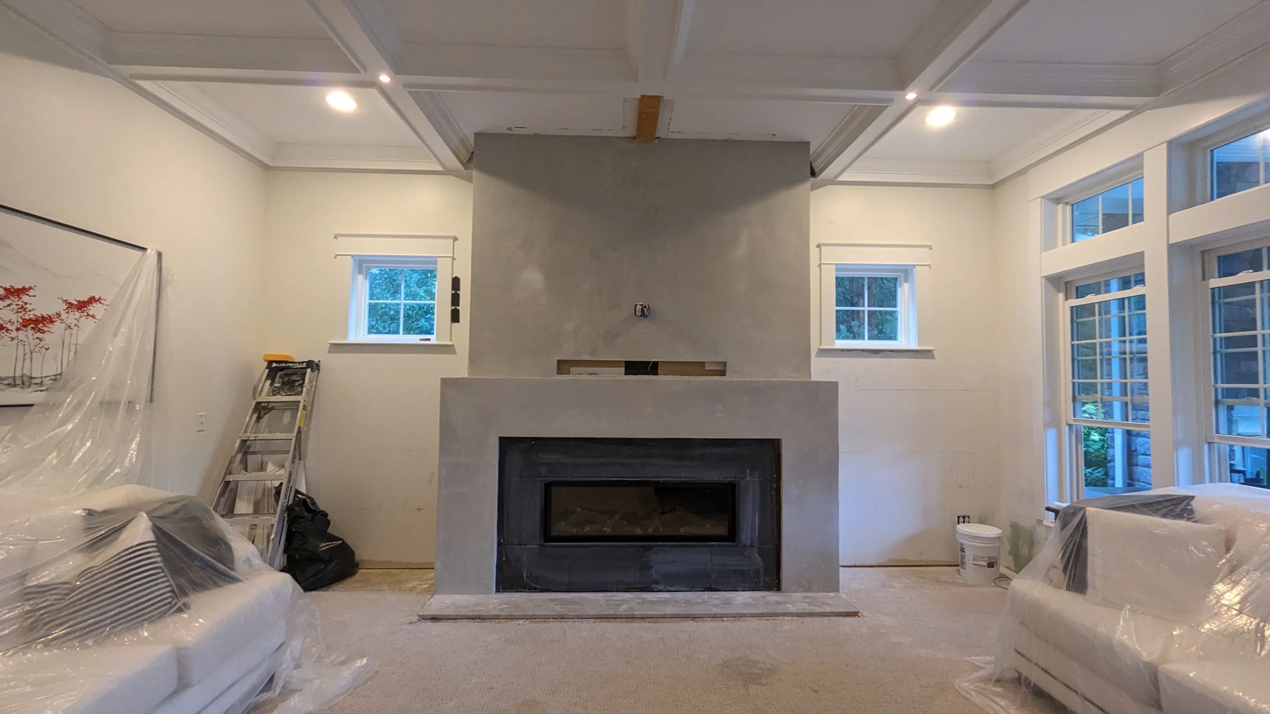 Living room under renovation with a marble fireplace, large windows, plastic-covered furniture, a ladder, and construction materials.