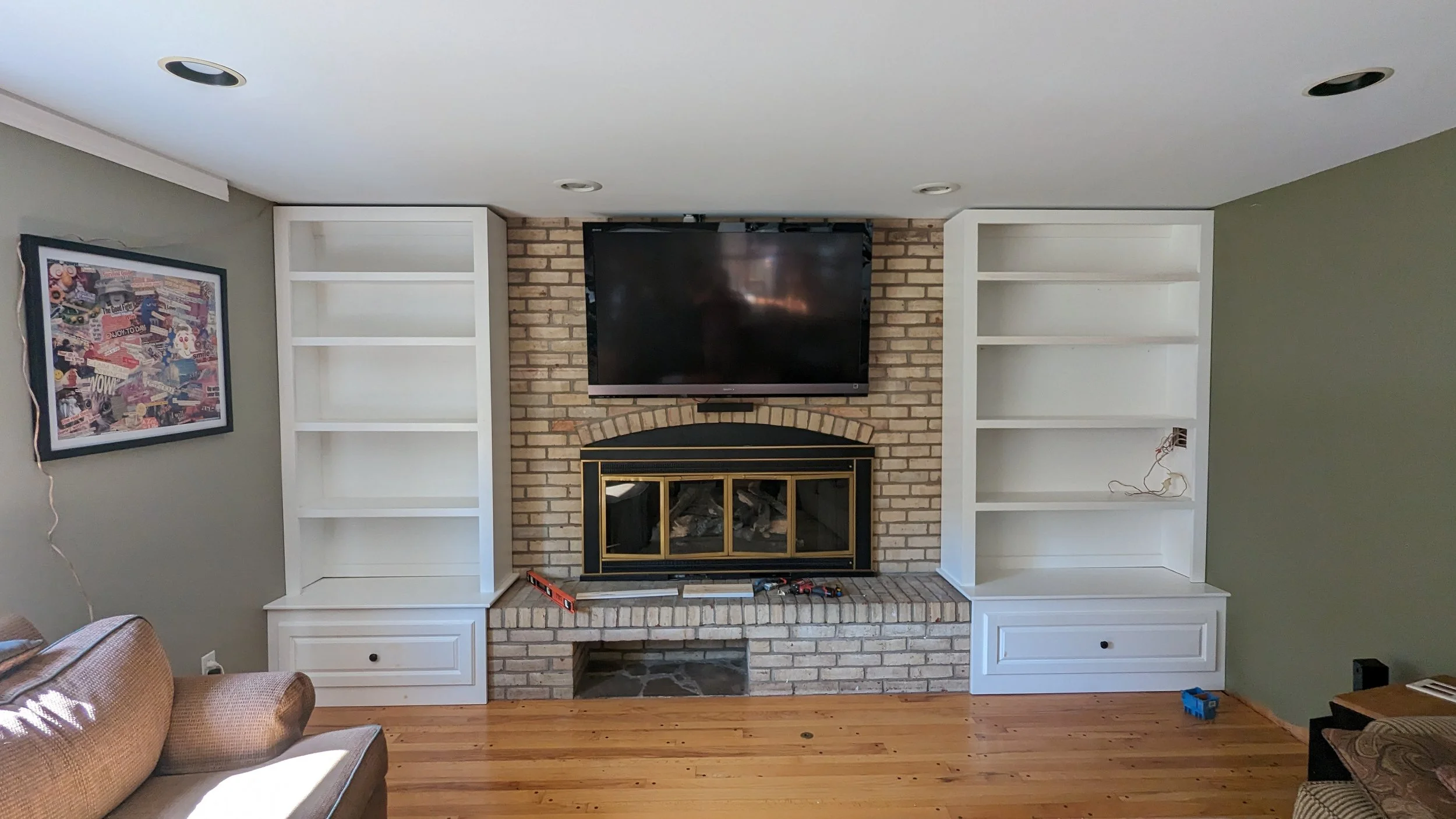 Living room with a fireplace, two white built-in bookshelves, a flat-screen TV mounted above the fireplace, and a framed collage on the wall to the left. There is a beige sofa on the left, and the room has wooden flooring and a green-painted wall.