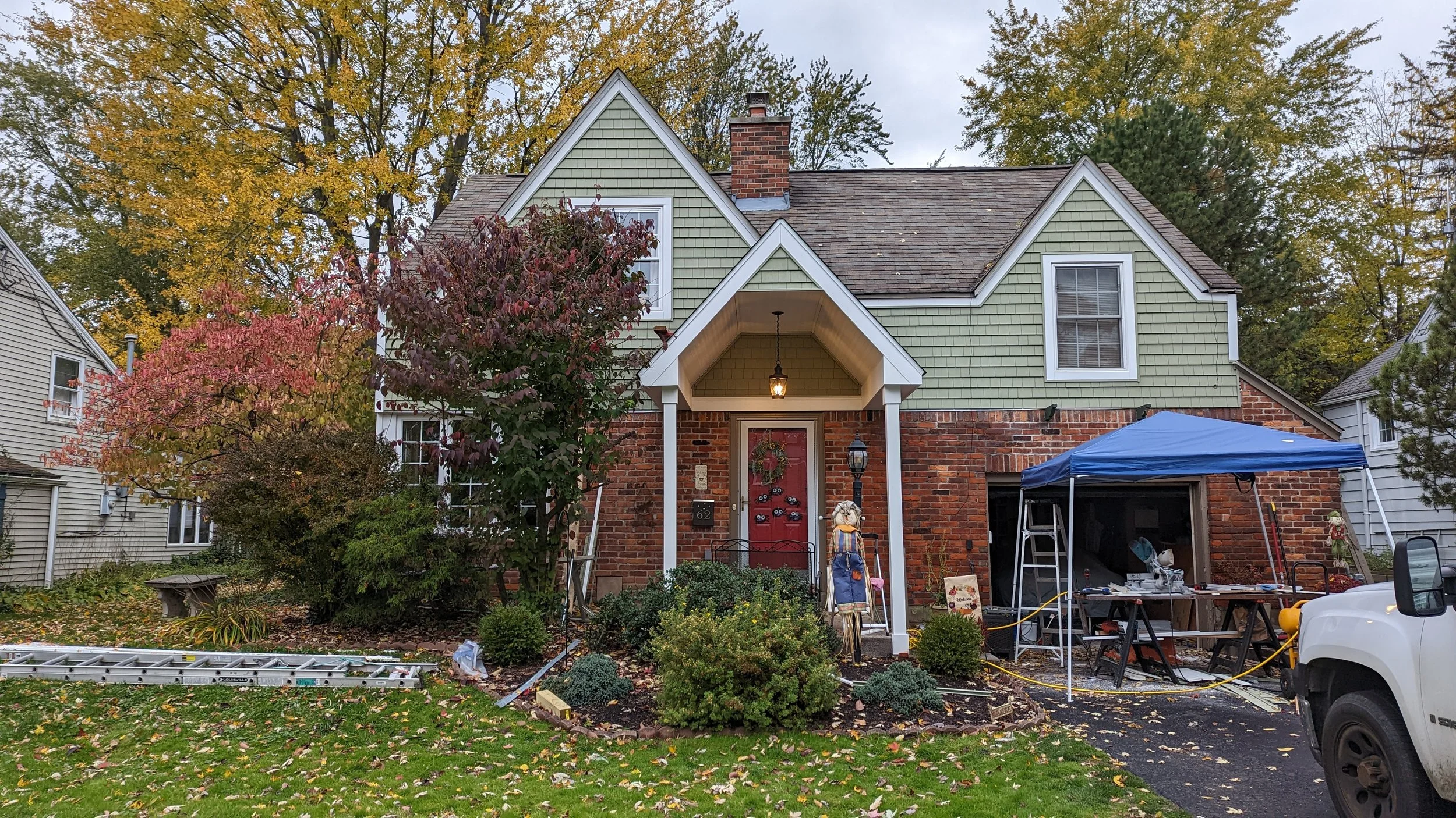 Front of a house with a brick lower level and green siding upper level, red front door with wreath, and trees with fall foliage. There is a blue canopy tent, outdoor decorations, and tools near the driveway.