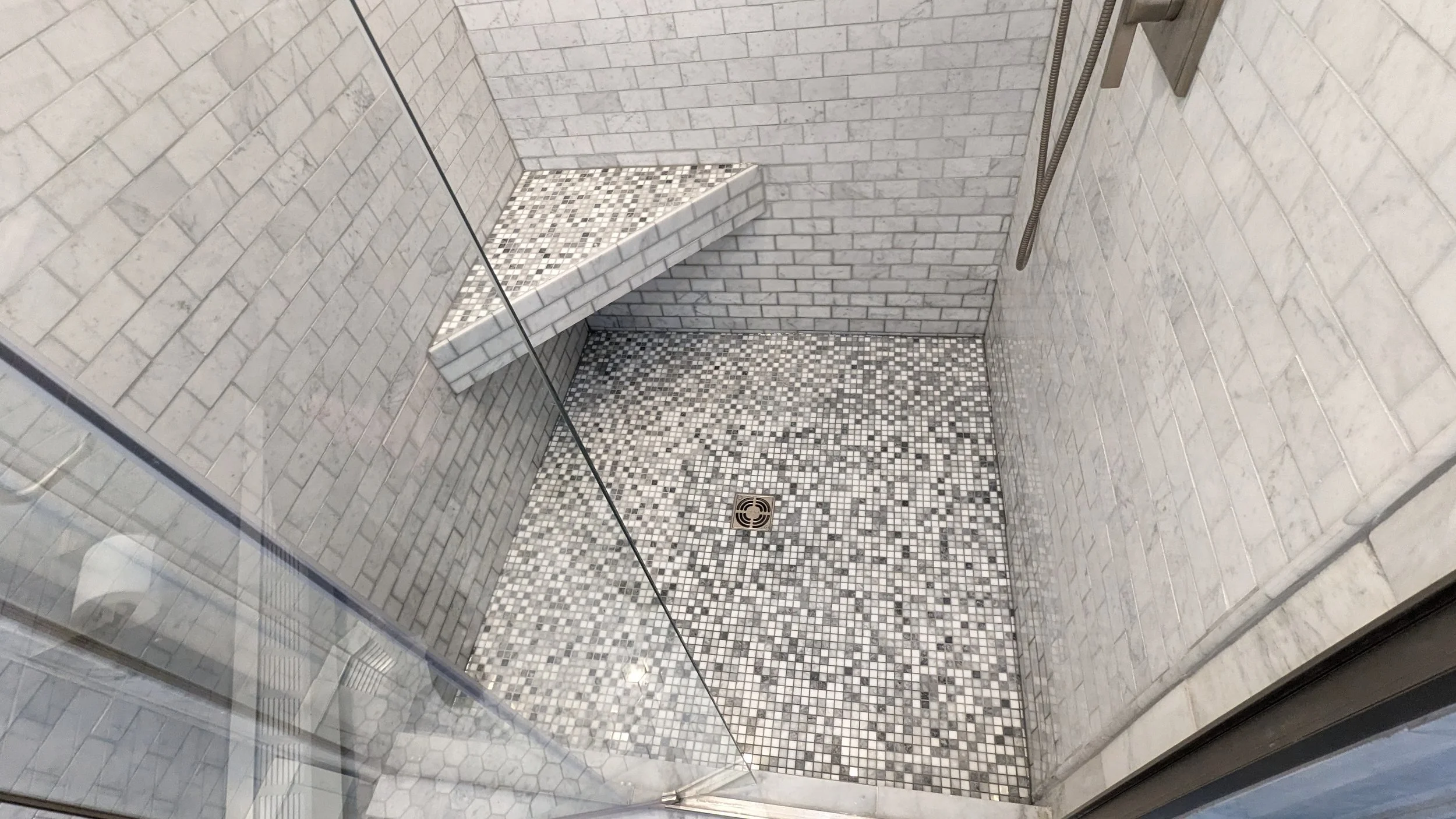 Inside a modern walk-in shower with white subway tile walls, a built-in corner shelf with small mosaic tiles, a floor drain, and a glass enclosure.