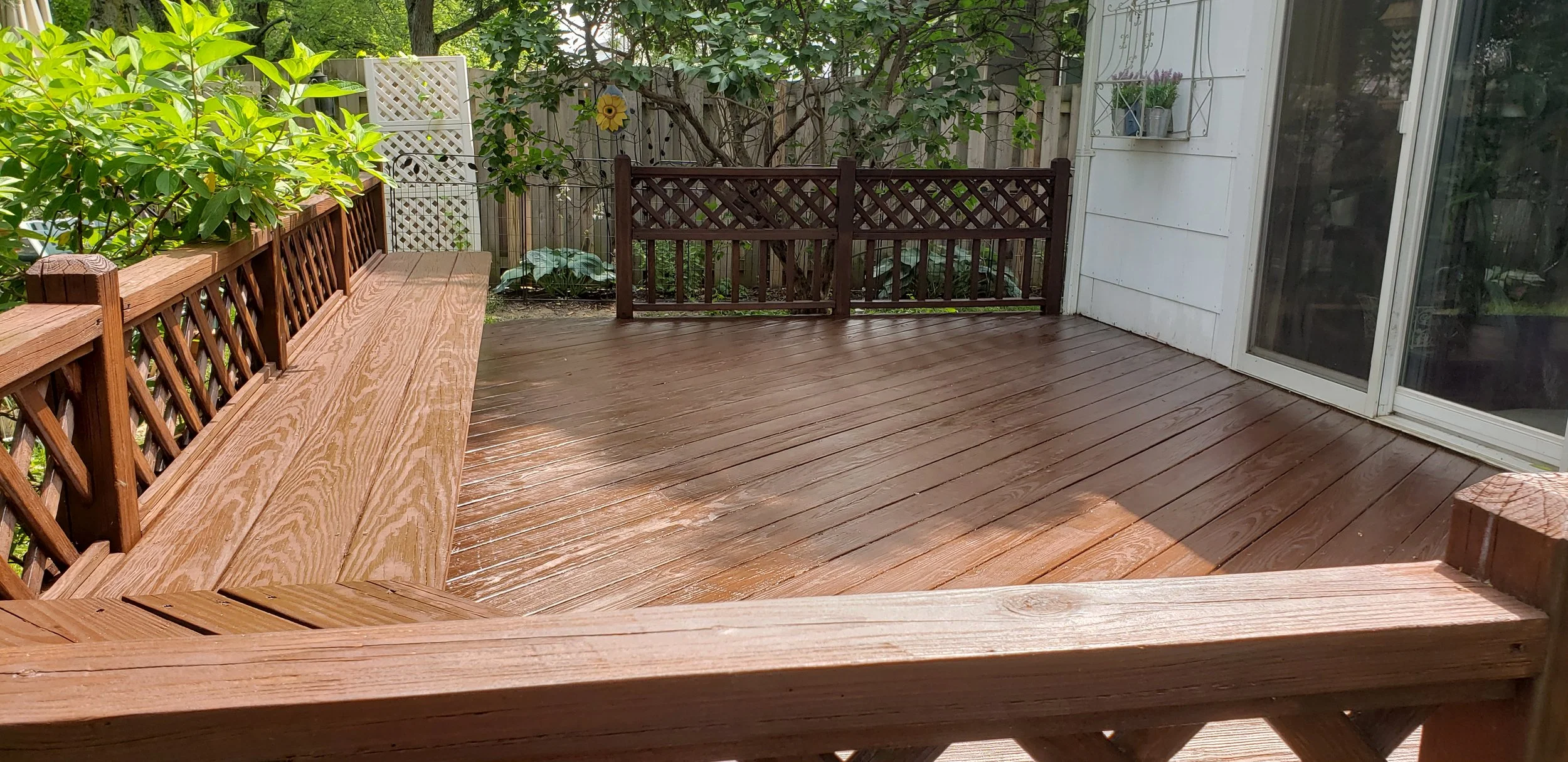 A wooden outdoor deck with a bench on one side, surrounded by greenery and a wooden fence. There is a sliding glass door leading into the house.